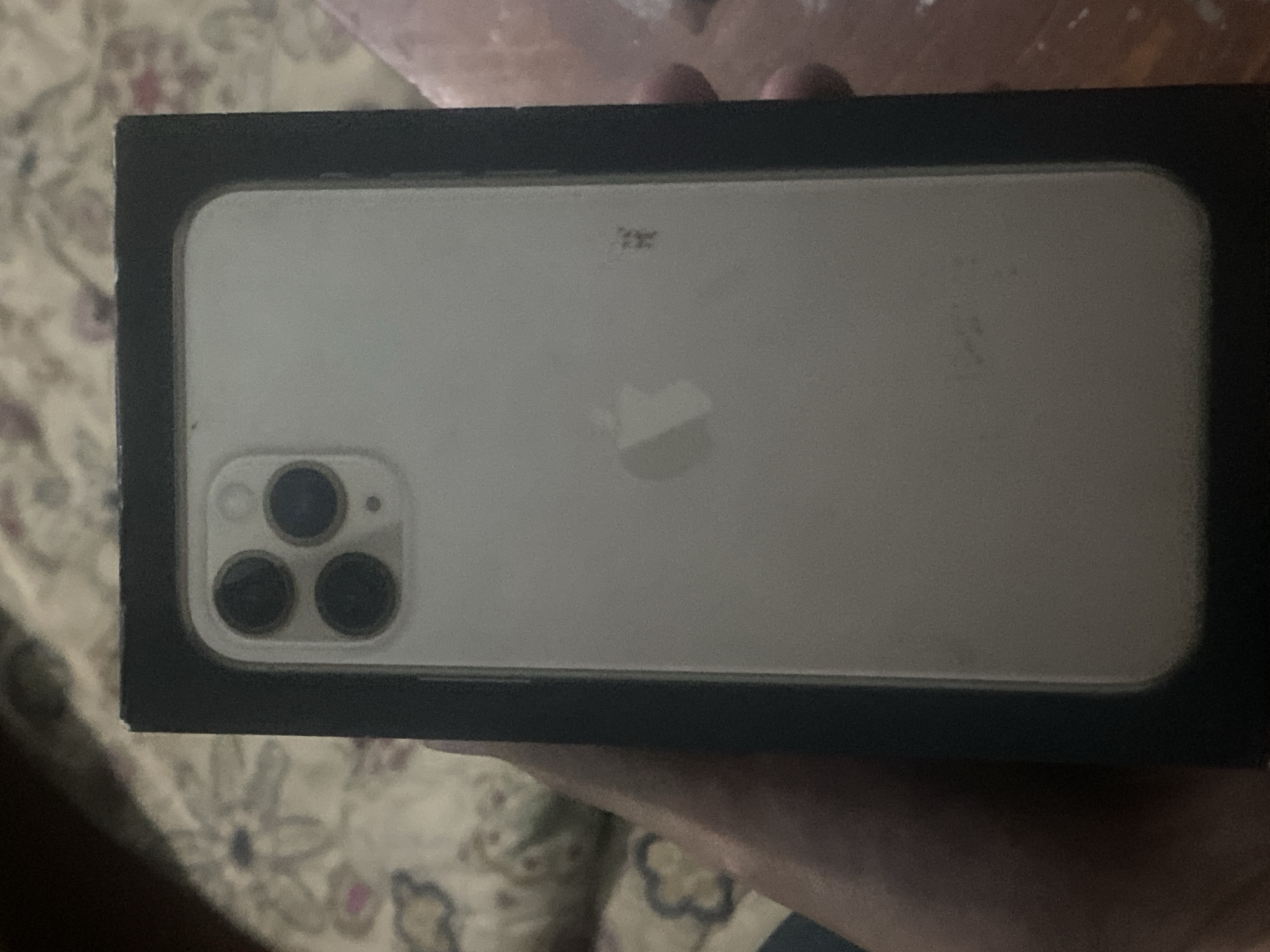 The image shows the back of a silver iPhone 11 Pro Max, which is partially visible within its black box packaging. The phone has a distinctive triple-camera system on the top left corner of its back panel. The Apple logo is visible in the center of the phone's back. The phone is held by a person whose hand is visible on the left side of the image. The background appears to be a patterned fabric, possibly a bedspread or blanket. The overall lighting is dim. The image was taken in the city of Dusti, Tajikistan.