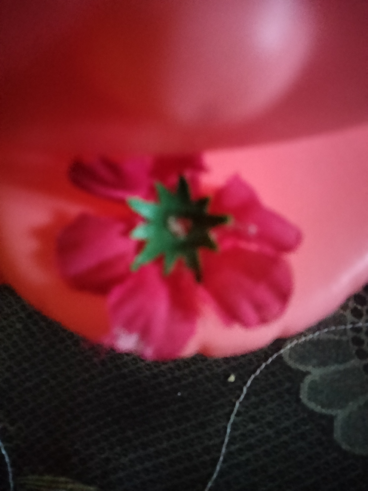 The image shows a close-up, blurred view of a pink flower with a green center, resting on a red surface. The red surface appears to be part of a larger object, possibly a bowl or a hat, with a lighter pink area suggesting a highlight or a different material. Below the red surface, a dark, textured material, likely fabric with a woven pattern, is visible. A thin white thread or string is also present on this dark fabric. The overall lighting is warm and diffused. There are no people or specific landmarks visible that would indicate a location in Payagaboh, Indonesia.