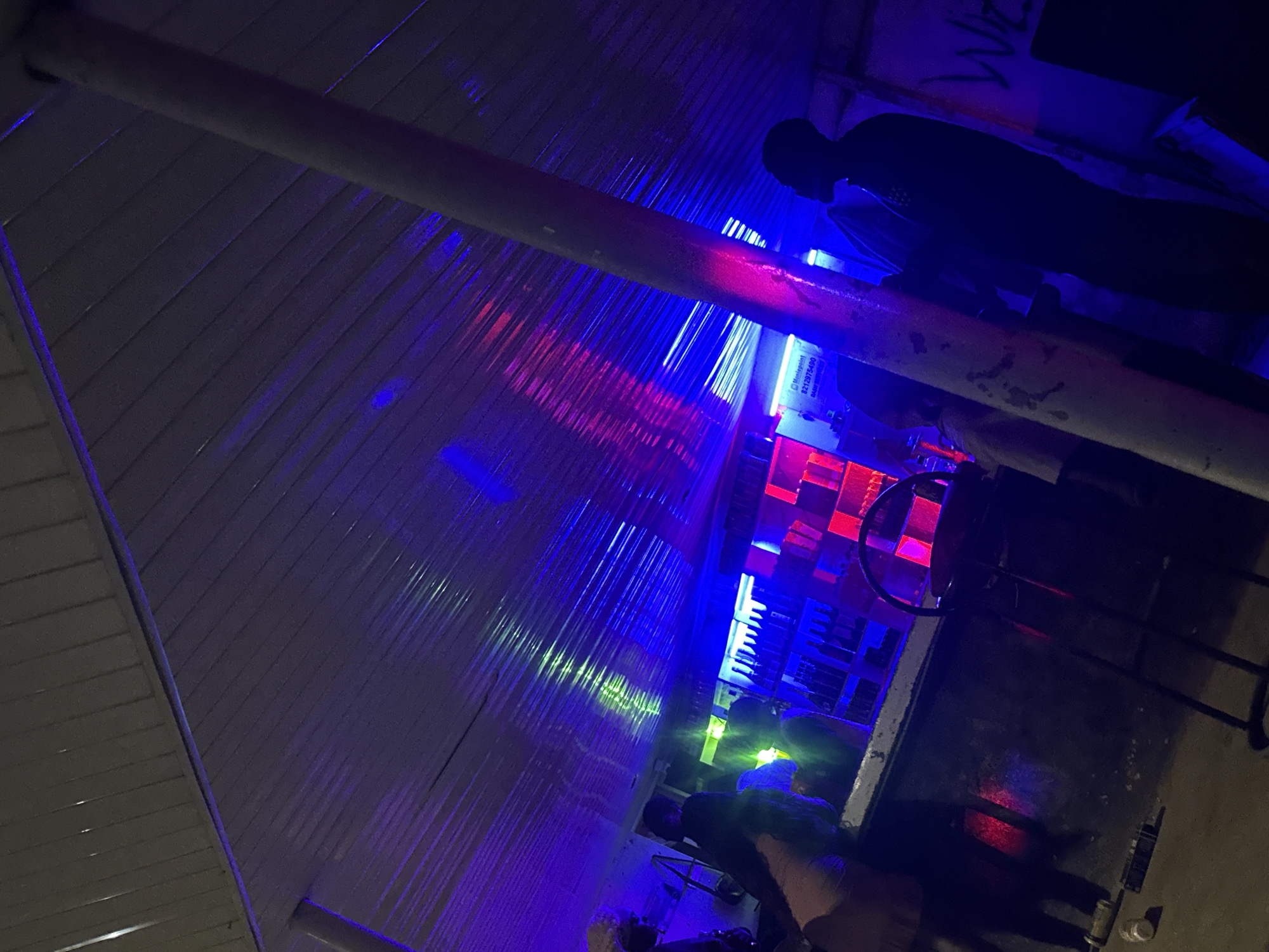 The image depicts an indoor bar scene illuminated by vibrant blue and pink neon lights, casting colorful reflections on the corrugated metal ceiling and surrounding surfaces. Several individuals are present, though their features are obscured by the low lighting and silhouettes. In the background, shelves stocked with bottles are visible behind a counter, suggesting it is a functional bar. A stool is positioned near the counter. Graffiti-like text, including "WE", is present on a wall to the right. The overall atmosphere suggests a nighttime setting within a bar or lounge, possibly located in Wumat, Nigeria, given the context provided. The scene is dimly lit, with the neon lights creating a dynamic and somewhat abstract visual effect.