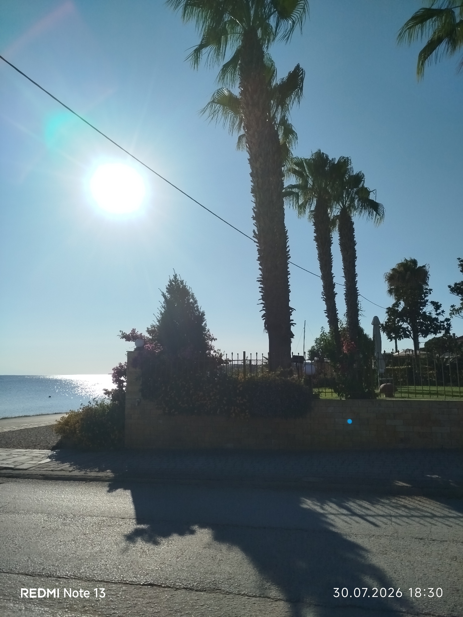 The image depicts a sunny day scene, likely in a coastal area of Sithonia Municipal Unit, Greece, given the presence of palm trees and the sea. The sun is bright and high in a clear blue sky, casting a strong glare. In the foreground, a paved road surface is visible, with shadows of trees and possibly a fence cast upon it. A stone wall runs horizontally across the middle ground, with dense green foliage and some flowering plants growing over it. Behind the wall, several tall palm trees are prominent against the sky. Beyond the wall and foliage, the shimmering surface of the sea can be seen, with a narrow strip of beach in the immediate foreground of the water. The overall atmosphere is bright and warm, suggesting a summer day. A watermark at the bottom left reads "REDMI Note 13", and at the bottom right, "30.07.2026 18:30" indicating the date and time the photo was taken. There are no people or animals visible in the image.