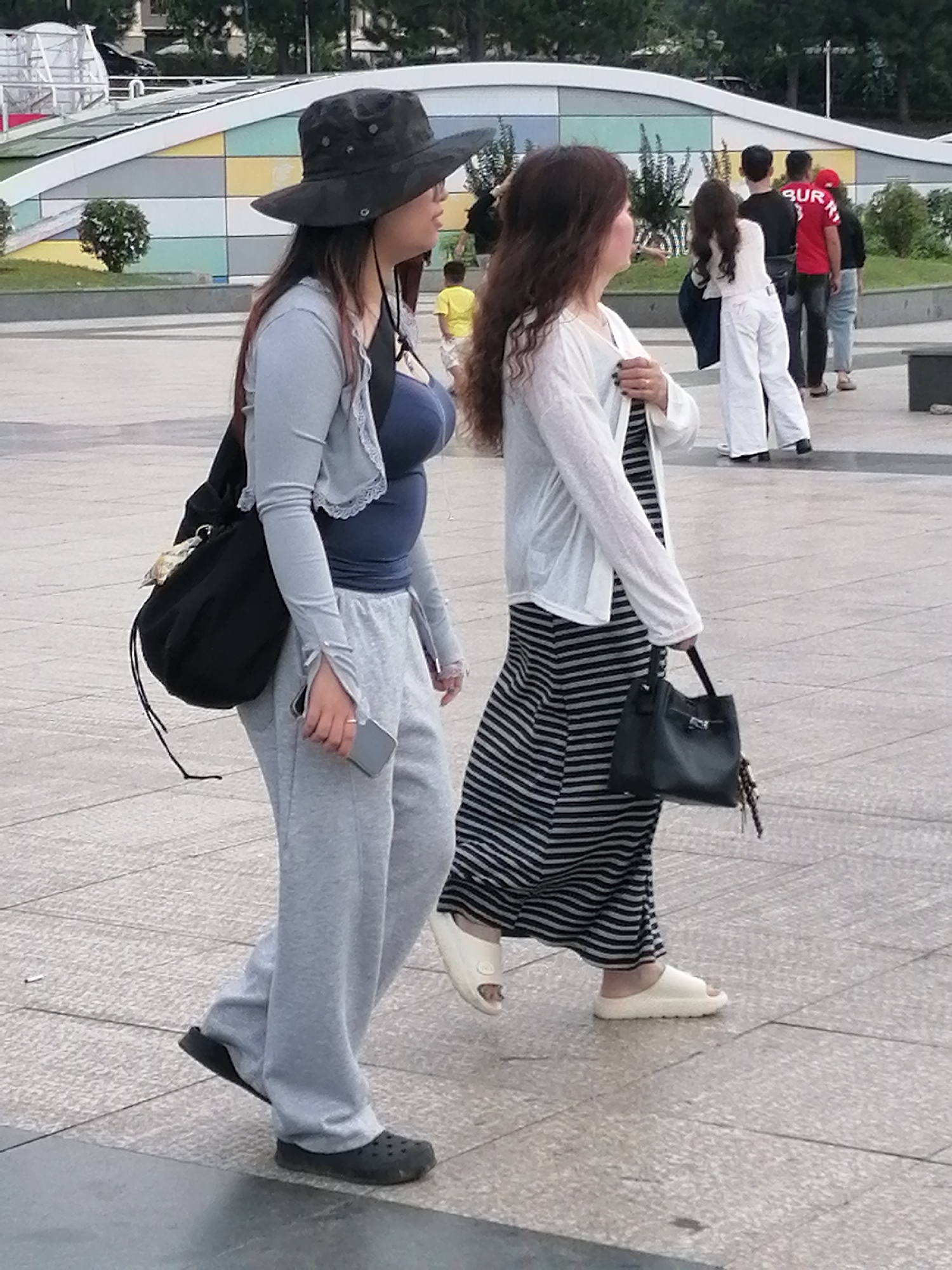 The image depicts an outdoor scene, likely a public square or walkway, with paved ground and a colorful, abstract architectural structure in the background. Two women are walking from left to right in the foreground. The woman on the left wears a wide-brimmed dark hat, a light gray long-sleeved top over a dark blue top, and light gray sweatpants. She carries a large black bag over her left shoulder and holds a silver mobile phone in her right hand. She is wearing dark clogs. The woman on the right is wearing a white open-fronted cardigan over a black and white striped maxi dress. She is carrying a black handbag in her right hand and wearing white slides. In the background, a group of people are walking away from the camera, including a person in a red shirt with "BUR" visible, and a person in a red hat. A young boy in a yellow shirt is also visible in the mid-ground. The environment appears to be a park or plaza, with some greenery visible near the background structure. The lighting suggests it is daytime. There is no specific indication of the location being Da Lat, Vietnam within the image itself.