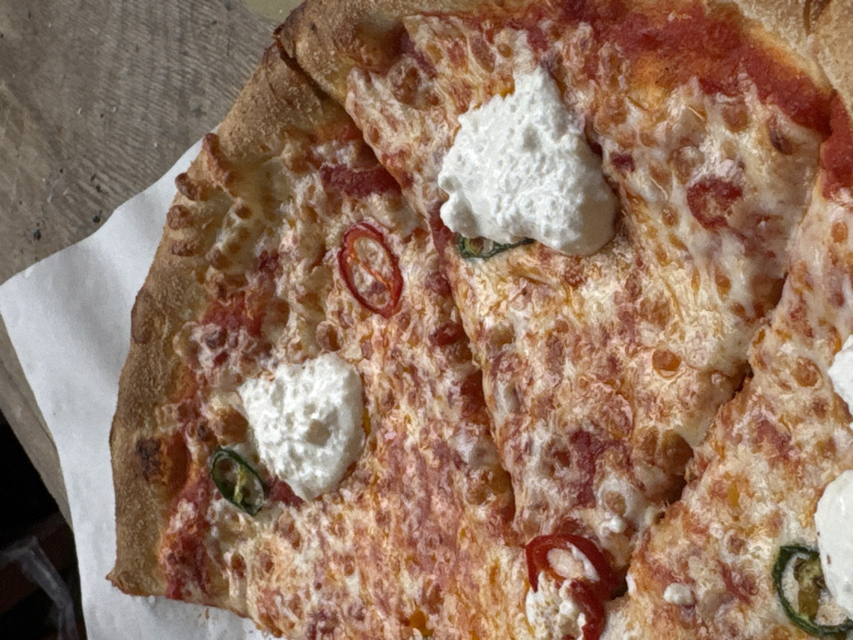The image displays a close-up, top-down view of a pizza cut into multiple slices. The pizza features a base of red tomato sauce and melted, browned cheese. Several distinct dollops of white, creamy cheese are visible across the slices. Scattered on the pizza are thin, circular slices of both red chili peppers and green chili peppers. The pizza rests on a piece of white paper, which lies on a textured, light brown surface, likely wood, exhibiting grain patterns. No individuals or active interactions are visible. The scene is located in Yerevan, Armenia.