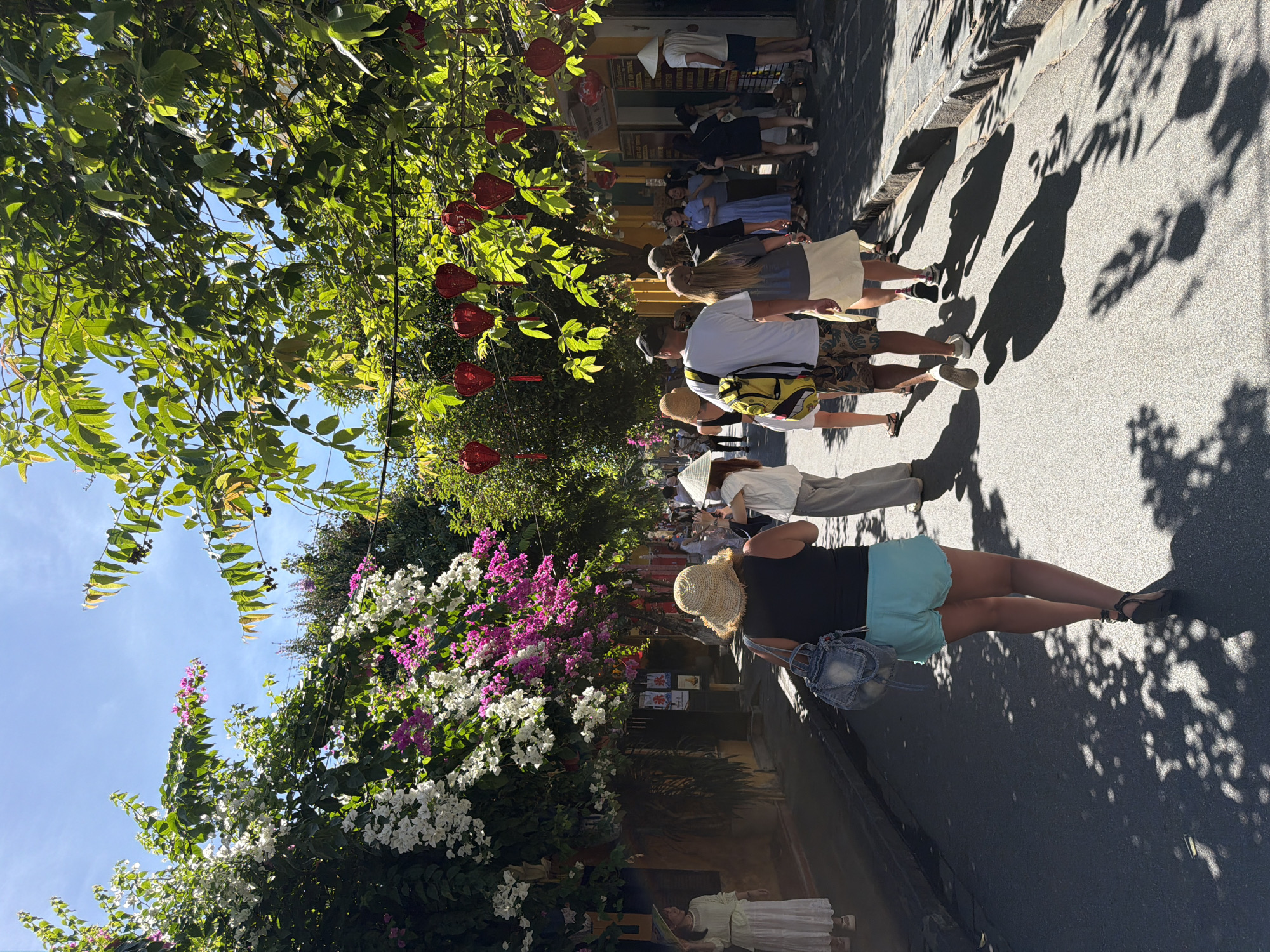 In Hội An Ward, Vietnam, a sun-drenched street is lined with vibrant bougainvillea in shades of white and pink, casting dappled shadows on the asphalt. A group of tourists walks down the street. In the foreground, a woman wearing a straw hat and turquoise shorts walks away from the camera, carrying a denim backpack. To her right, a man in a white t-shirt and patterned shorts walks alongside a woman in a light-colored skirt. Other individuals, some wearing conical hats, are visible further down the street, browsing shops and walking. Red paper lanterns hang from trees overhead, adding a festive touch to the scene. The architecture on the left suggests historic buildings with yellow facades.
