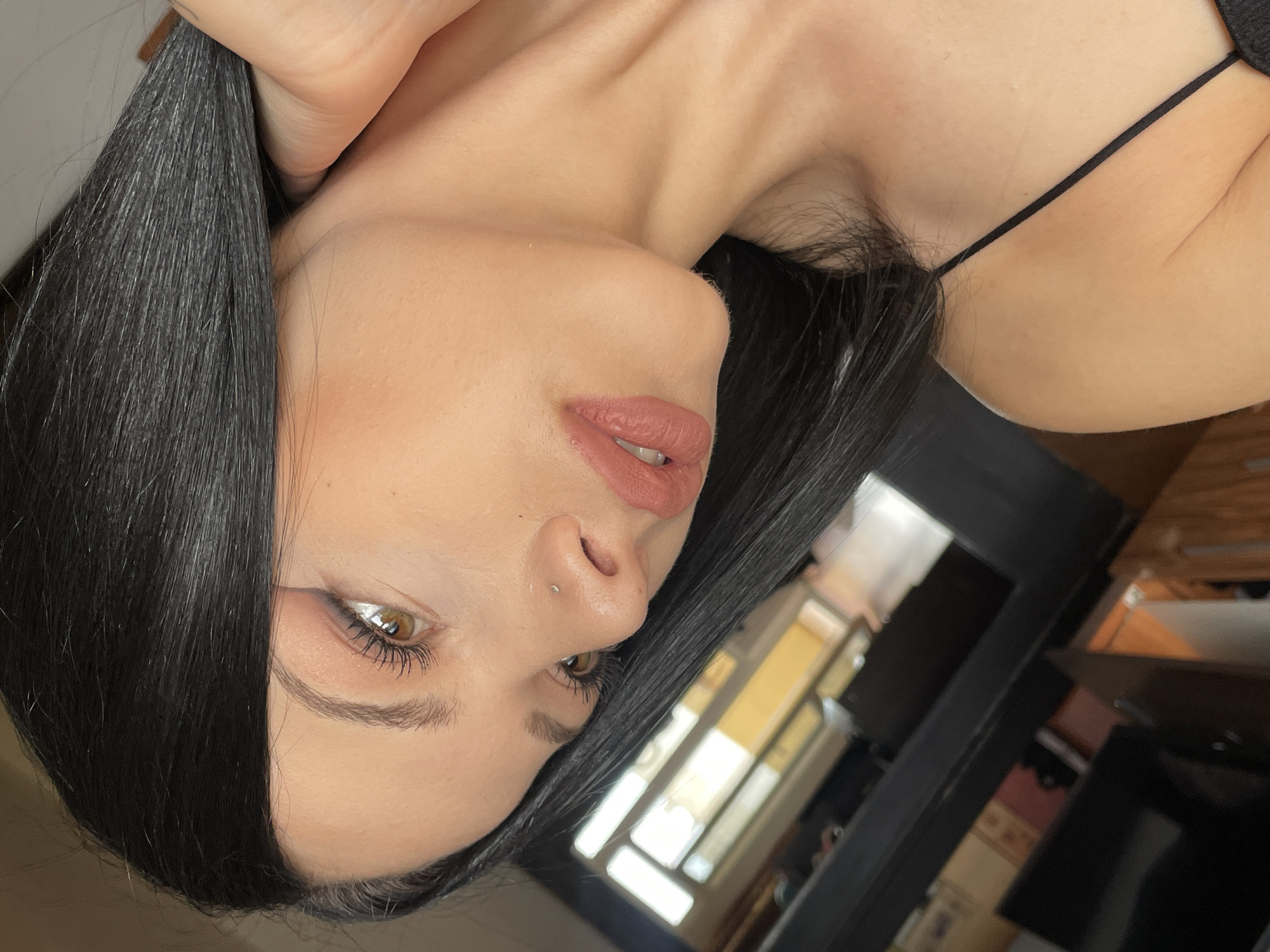 A young woman with long, dark hair is shown in a close-up, side-profile shot. She is wearing subtle makeup, including eyeshadow and lipstick. A small nose stud is visible. The background is blurred, but appears to be an indoor setting with furniture and possibly a window reflecting light. There is no discernible indication of the specific location within Stara Zagora, Bulgaria. The image focuses on her facial features and upper body.