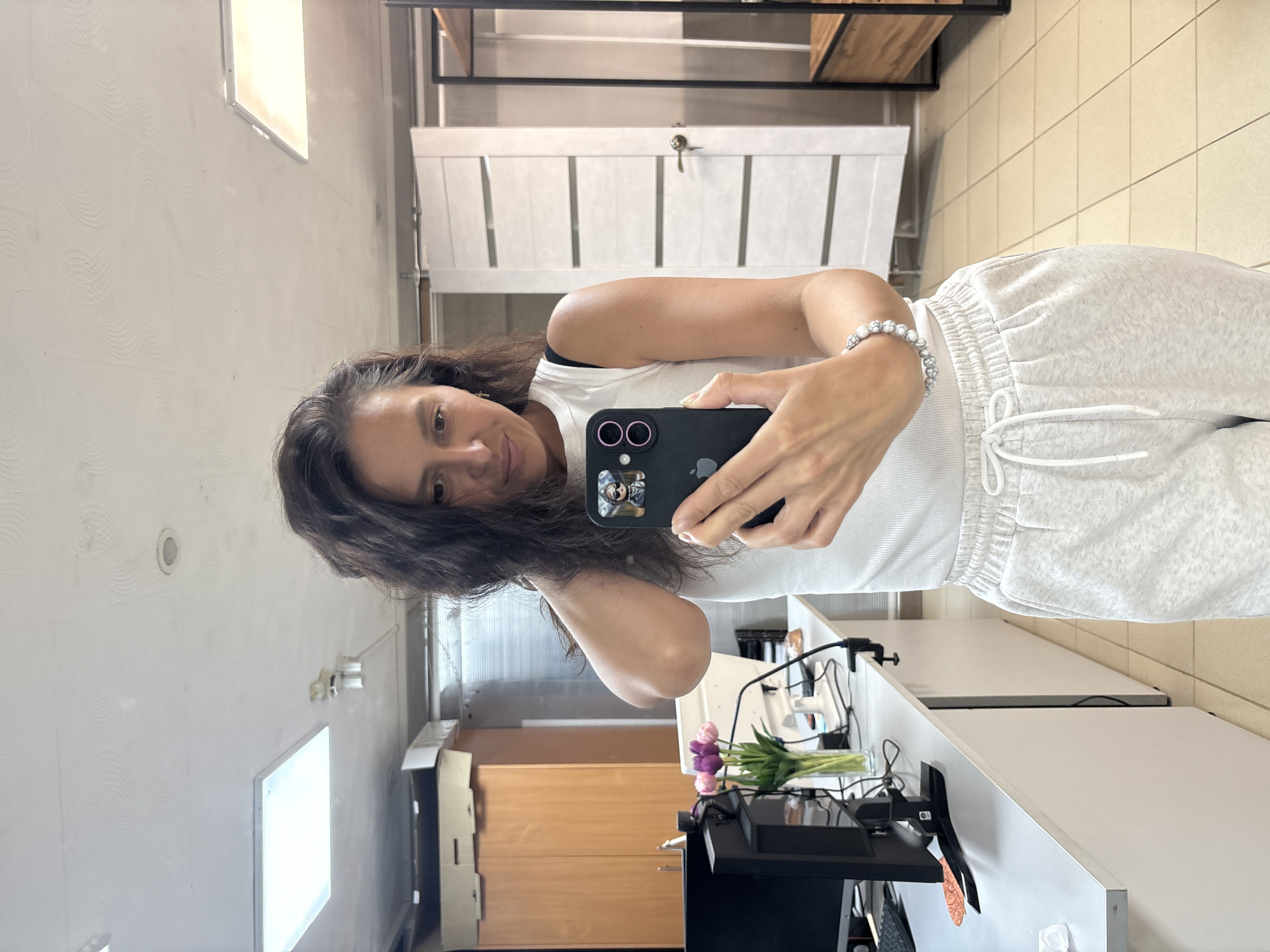A person is taking a selfie indoors. The person has dark hair and is wearing a white tank top and gray sweatpants. They are holding a black iPhone in their right hand, which has a sticker on it. A white bracelet is on their right wrist. Behind the person is a white door with horizontal panels and a shelving unit. A desk with a monitor, keyboard, and flowers is also visible. A window and cabinets are visible. The location may be in Chaikovsky, Russia.
