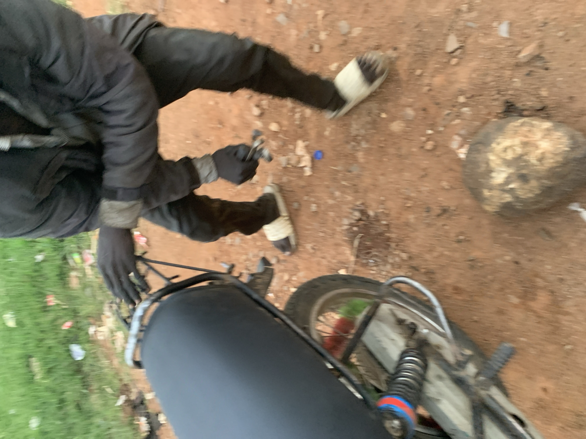 A person wearing a black jacket, black pants, and gloves is working on the rear section of a motorcycle in Barakin Lerawi, Nigeria. The motorcycle has a black seat and a visible rear suspension with red and blue accents. The ground is a mix of red dirt and gravel, with some green grass and scattered debris in the background. The person is holding a tool in their gloved hands, appearing to be engaged in maintenance or repair work. The overall scene suggests an outdoor, possibly informal, setting.