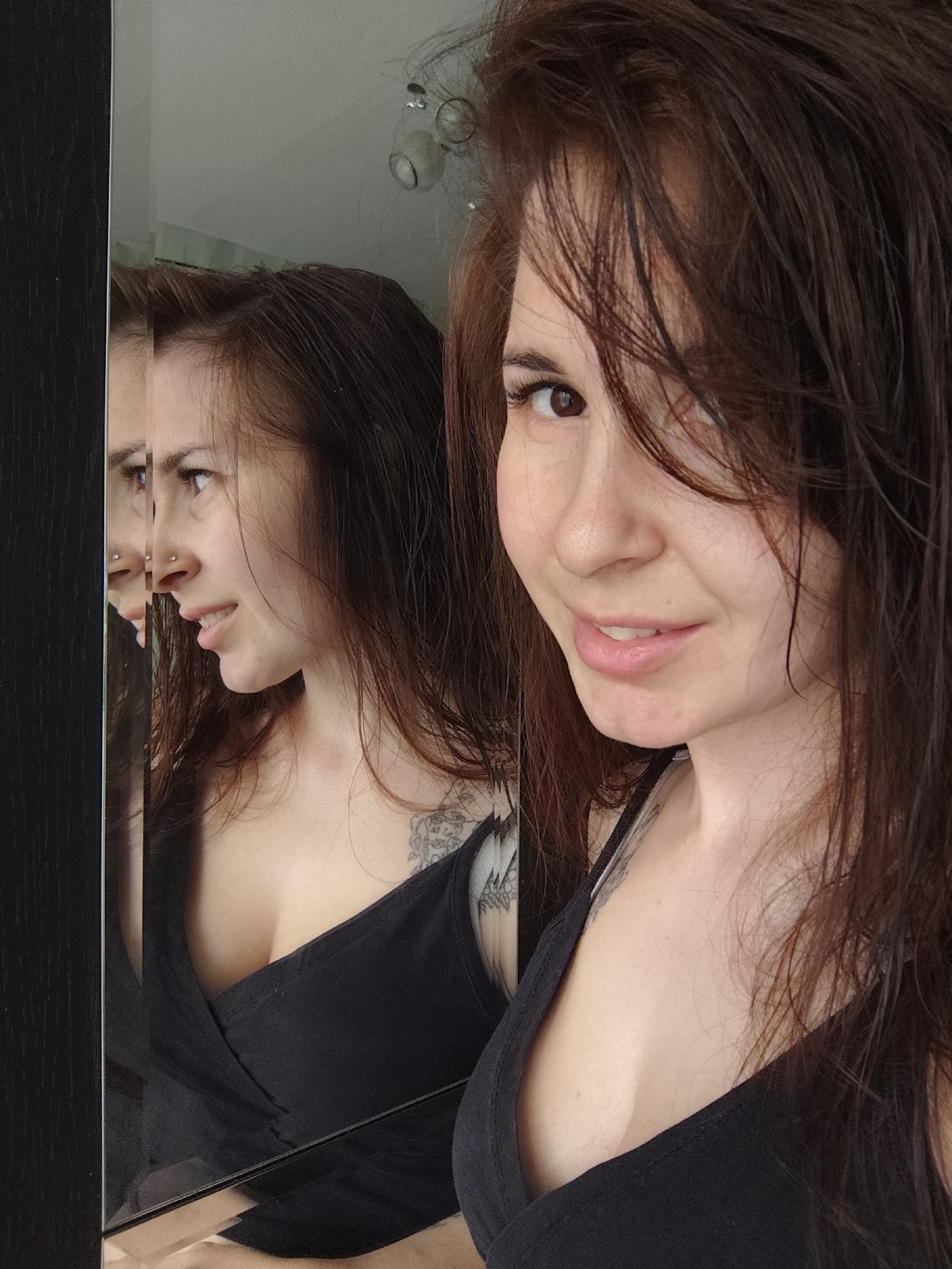 This image captures a young woman taking a mirror selfie in an indoor setting, likely in Фоки, Russia. The main subject is a woman with long, dark brown, possibly damp hair, which falls across her face and shoulders. In the foreground, she is looking towards the viewer with a soft, slight smile.
To her left, her reflection is prominently visible in a large, vertically oriented mirror, revealing her profile as she looks away. This reflection clearly shows a small piercing on her left nostril and a tattoo on her right shoulder, detailed with intricate patterns. She is dressed in a black top, possibly a tank top or camisole with a wrap-style front.
The mirror itself has a dark, possibly wooden, frame along its left edge. The reflection also offers glimpses of the interior environment, including a light-colored wall and a ceiling light fixture at the top. The lighting suggests it is daytime. The woman's relaxed expression contributes to a calm and intimate atmosphere of a casual moment captured indoors. No visible text is present in the image.