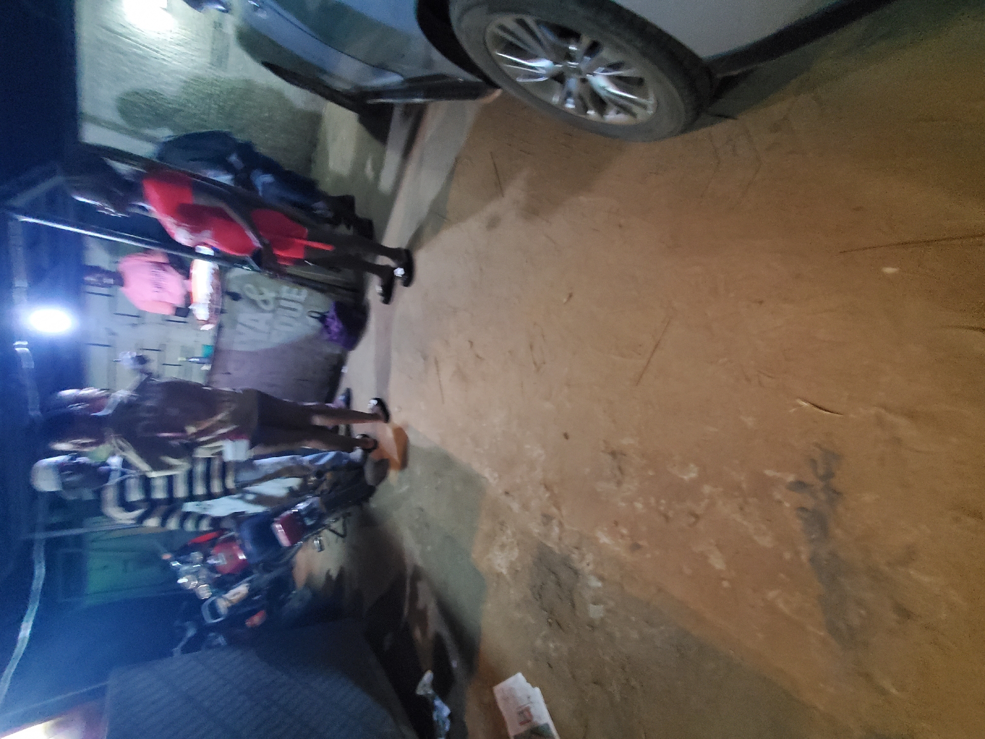 The image shows an outdoor scene in Nnewi, Nigeria, at night. The area is illuminated by a light source in the upper left of the image. A car is parked at an angle in the upper right of the image. There are at least two people visible. One individual wears a striped shirt and stands beside a motorcycle. Another person wears a red shirt and stands near the top of the image. The ground is a mix of dirt and concrete.
