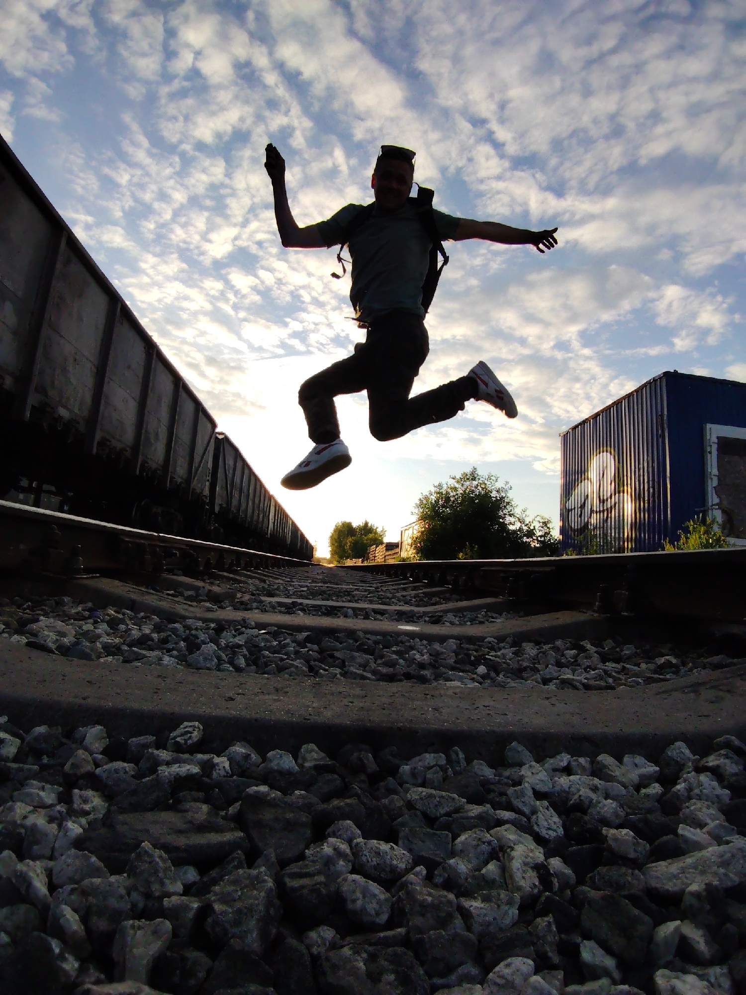 A low-angle photograph taken in Chaikovsky, Russia, depicts a male figure in mid-air over train tracks, silhouetted against a bright, partly cloudy sky. The individual wears a dark t-shirt, dark pants, white sneakers with red accents, and a dark backpack. His right arm is raised with a clenched hand, while his left arm is extended horizontally. Sunglasses are positioned on top of his head.
The sky is blue with scattered white altocumulus clouds, and a strong backlighting effect emanates from a bright horizon line on the right, indicating a low sun. The foreground features multiple sets of train tracks, consisting of steel rails, sleepers, and grey ballast rocks. A long line of dark freight train cars occupies the left side, extending into the background. On the far right, a blue corrugated metal container or small building, adorned with white graffiti and featuring a boarded-up opening, stands beyond the tracks. Green trees and bushes are visible in the middle ground, further along the tracks, also silhouetted by the backlight.