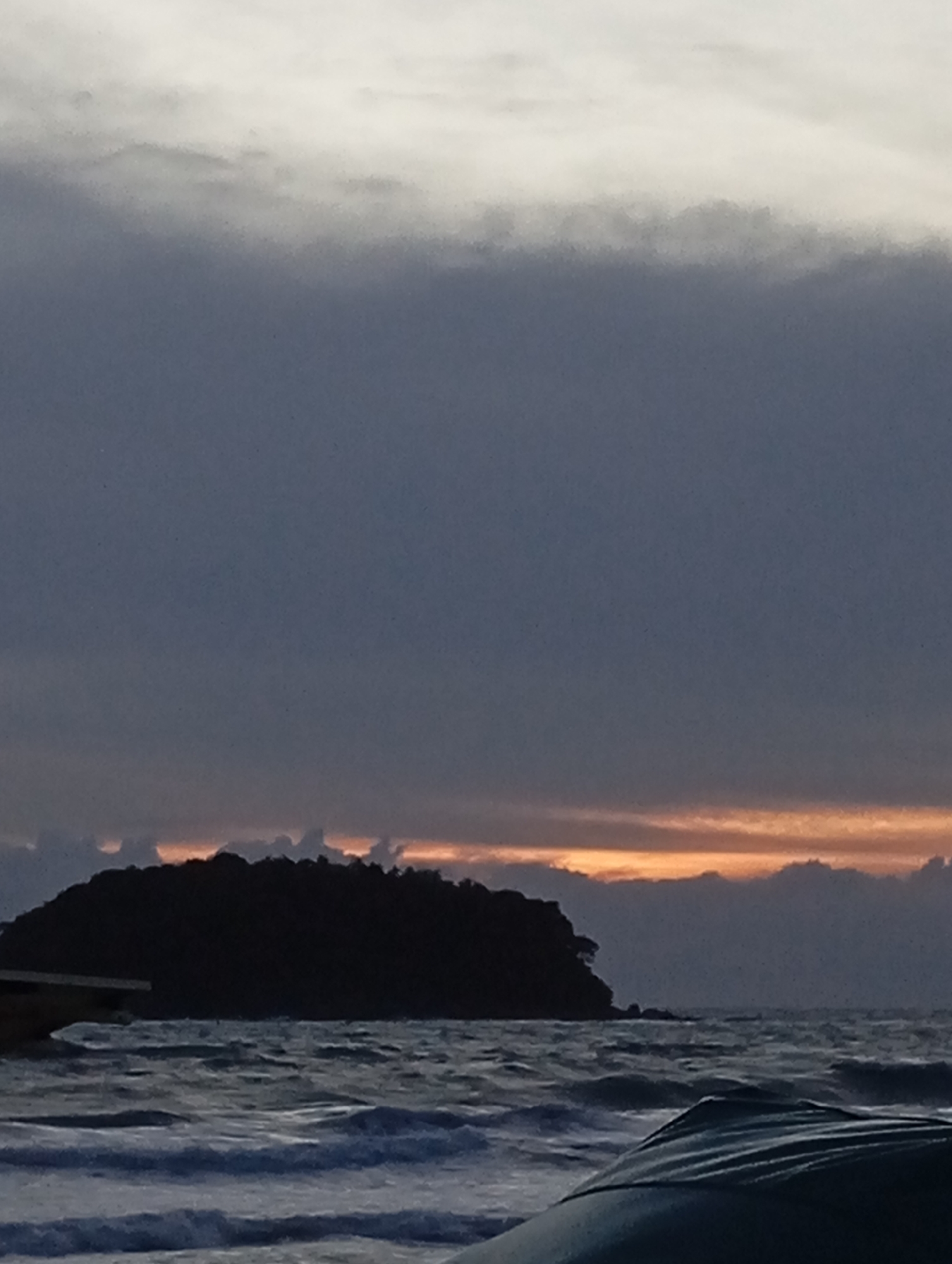 A silhouetted island sits on the horizon in Karon, Thailand, bathed in the muted light of dusk. The sky is a gradient of dark grays and blues, with a faint band of orange glow just above the island, suggesting the last rays of sunlight. The sea is choppy, with whitecaps indicating moderate waves. The foreground shows the dark, curved edge of what appears to be a boat or raft. No people are visible in the image. The overall atmosphere is one of quietude and the transition from day to night over a tropical seascape.