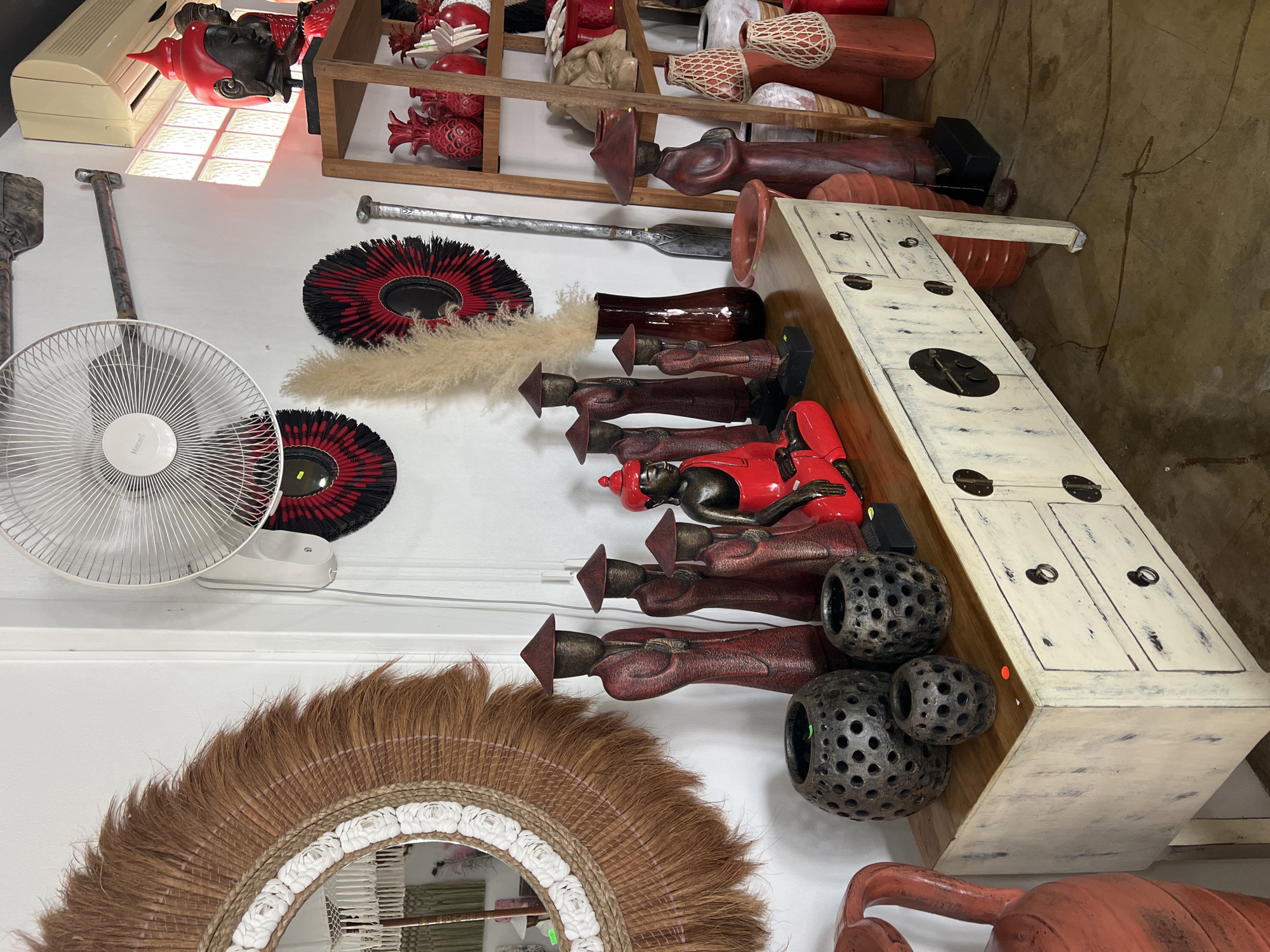 This indoor shot displays a variety of objects situated against a white wall and part of a weathered, textured beige floor. The scene appears to be inside a store or workshop, given the assortment of decorative items.
On the wall, a white oscillating fan is visible, along with a large circular mirror bordered with brown and white decorations. Above the mirror, there are two decorative circular mats in red and black, and a bundle of beige-colored plumes or feathers.
A shelf unit holds a selection of red and white decorative objects. Several wooden sculptures with conical hats are present, interspersed with a red figurine. Additional pottery or vases in earthy tones are also on display.
A white painted cabinet with dark metal hardware sits on the floor, adjacent to a cluster of dark, spherical objects with circular cutouts. The floor is scuffed and marked, providing a contrast to the white wall.