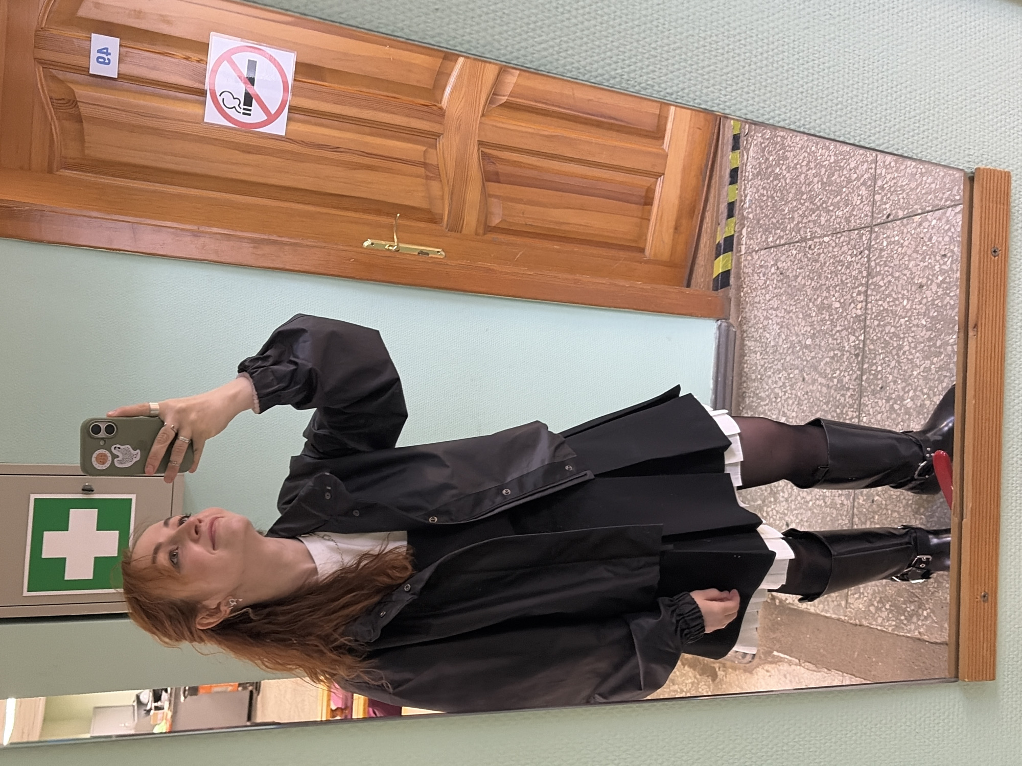 A person is reflected in a mirror, holding a cell phone in their left hand to take the photograph. The person has red hair and is wearing a black jacket and knee-high black boots. Behind the person in the reflection is a wall and a door. A "no smoking" sign is on the door. A green first-aid kit hangs on the wall. The location is in the city of Chaikovsky, Russia.