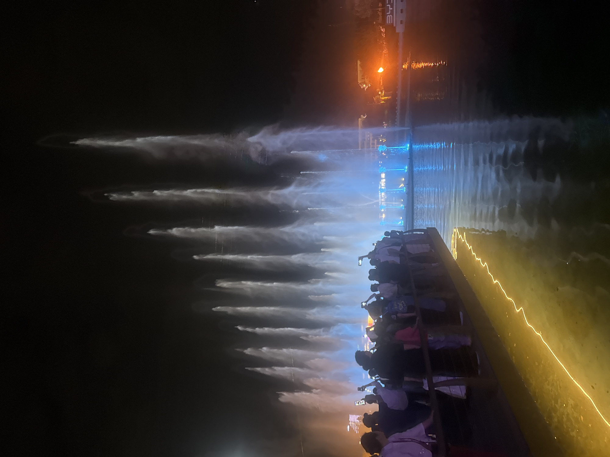 This nighttime image captures a bustling scene at the musical fountains in Magic City Park, Tashkent, Uzbekistan. A large crowd of people, primarily seen from behind or in profile, stands on a dark promenade in the foreground, observing a grand water display. Many individuals are holding up smartphones, actively recording or photographing the fountains.
The central and middle ground of the image is dominated by numerous powerful water jets erupting from a large body of water. These fountains are dynamically illuminated, predominantly with bright white and various shades of blue light, creating tall, fluid columns against the dark sky. The lights and the shapes of the water jets are clearly reflected on the calm surface of the water, enhancing the visual spectacle.
Along the edge of the promenade in the immediate foreground, a distinctive, undulating line of yellow light glows, casting a warm reflection onto the water. In the distant background, across the expanse of water, some indistinct buildings and structures are visible, faintly lit with warm orange and cool blue lights. To the far right, a white block-like structure with the letters "BYD" is partially discernible, indicating a specific brand presence within the park. The sky is dark and clear, accentuating the vibrant artificial lighting of the fountains and surrounding environment.