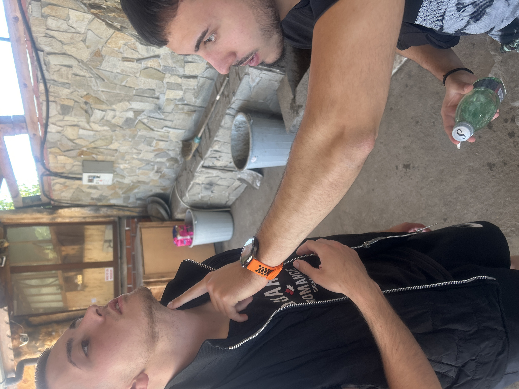 Two men are outdoors in what appears to be a courtyard or patio area. The location is Elhovo, Bulgaria. One man is lying on the ground wearing a black t-shirt and a black vest. He has a smartwatch with an orange band on his left wrist. The other man is standing, looking down at the man on the ground. He is holding a green bottle in his right hand. The courtyard has stone walls, a concrete floor, and several buckets are present. There is an electrical box on the wall. The scene suggests a moment of concern or assistance.