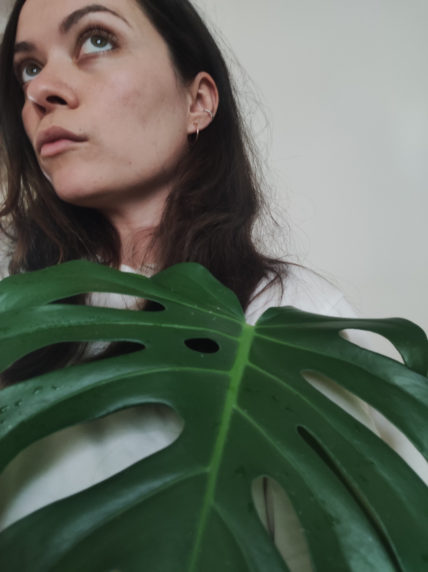 A young woman with dark hair and light skin is partially obscured by a large, green Monstera leaf. The woman is wearing a white shirt and has a nose ring and earrings. She is looking upwards and to the left. The background is a plain white wall. The image does not show Фоки, Russia.