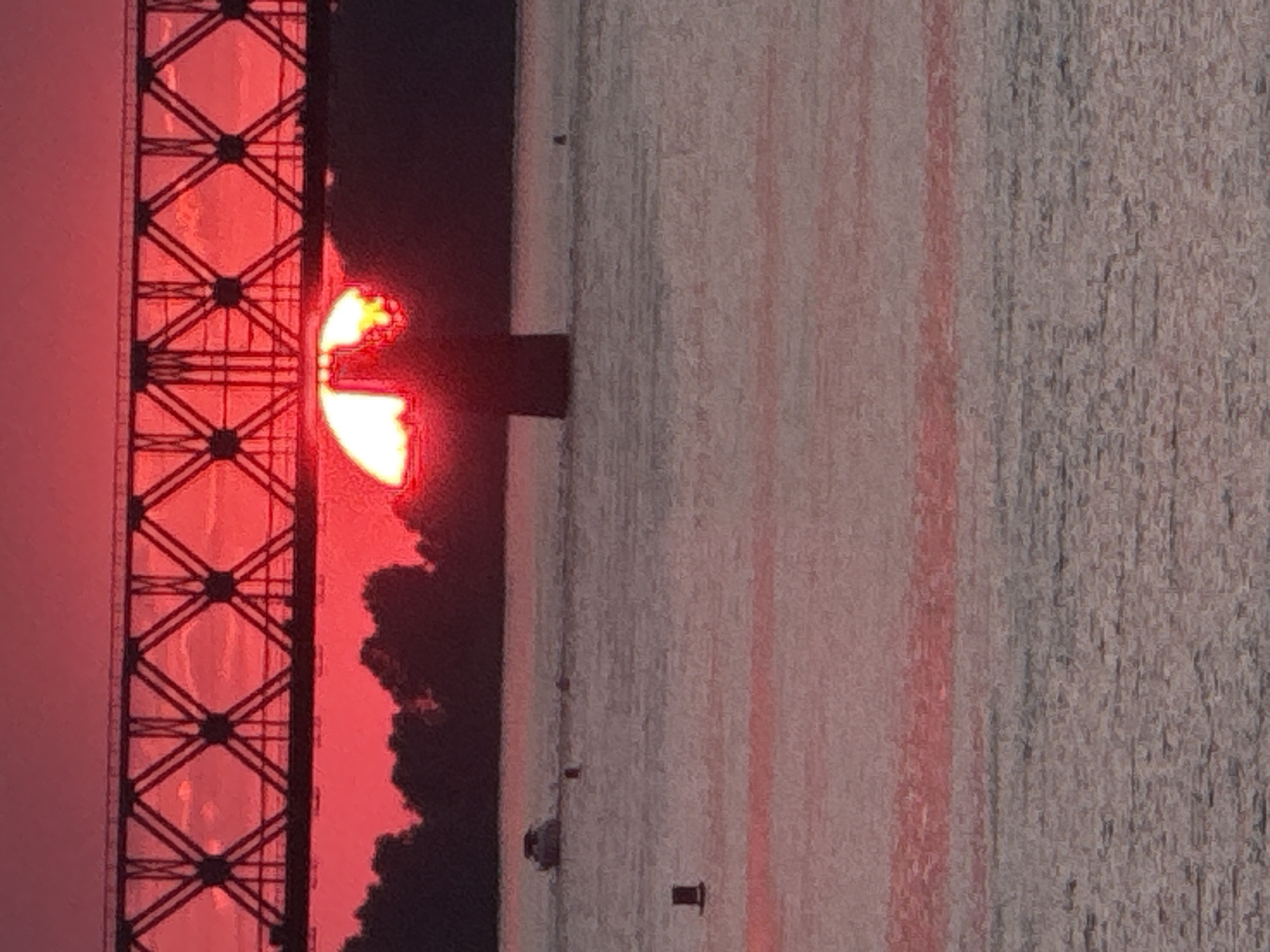 The image shows a sunset over a body of water, likely the Irtysh River, in Pavlodar, Kazakhstan. A bridge with a lattice structure spans across the top of the frame, with the setting sun partially visible behind one of its support piers. The sky is a vibrant red, indicating the time of day. In the water, a small boat is visible, along with several buoys. The trees and shoreline are silhouetted against the bright sky.