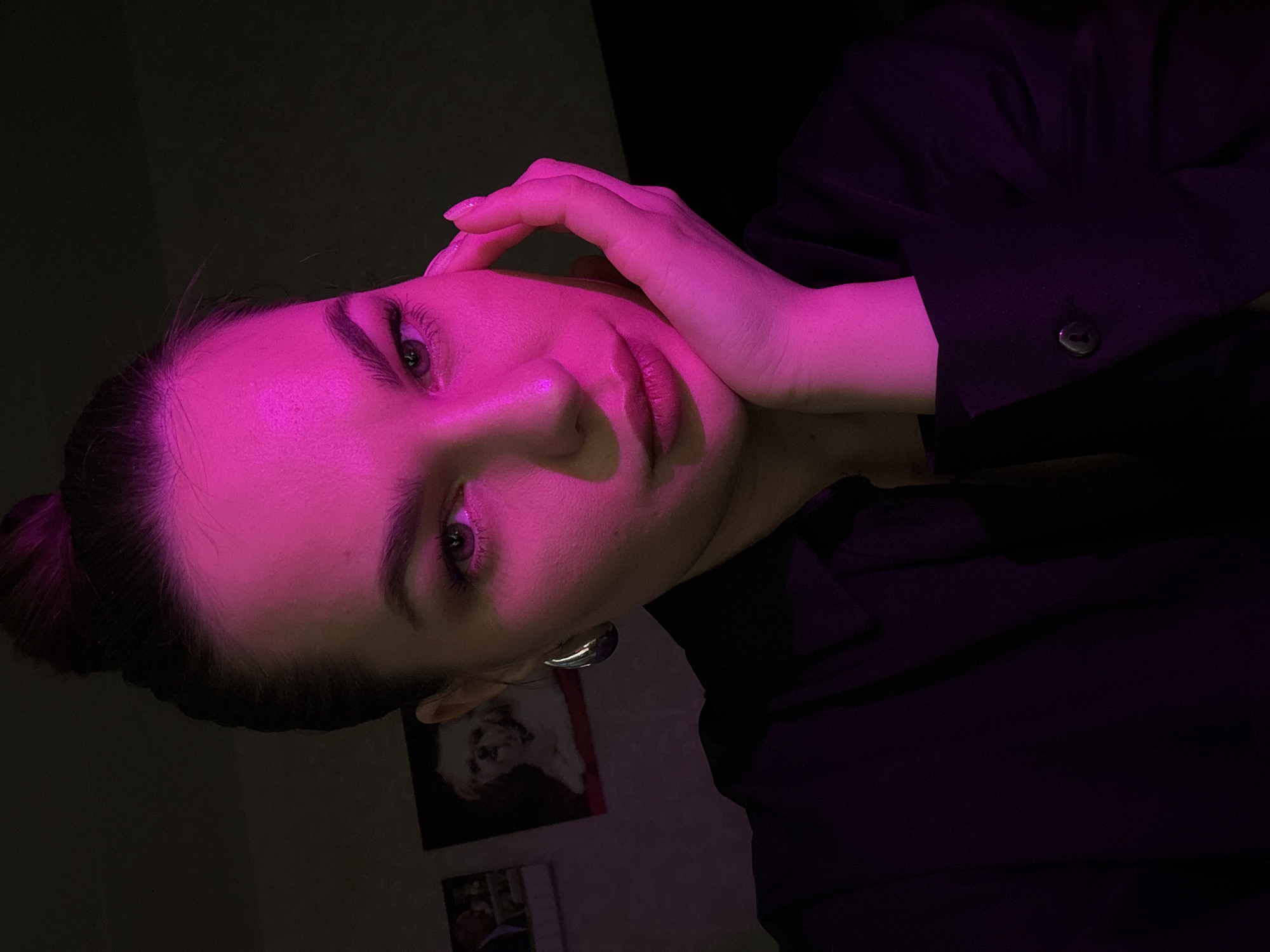 The image is a close-up selfie of a young woman with dark hair pulled into a bun. She is illuminated by pink lighting, which casts a vibrant glow on her face. Her left hand is resting on her cheek, and she is wearing a dark-colored shirt with a button. In the background, there are two framed pictures on a wall, one appearing to be of a dog and the other with people. The overall environment appears to be an indoor setting, possibly a room in Chaikovsky, Russia.