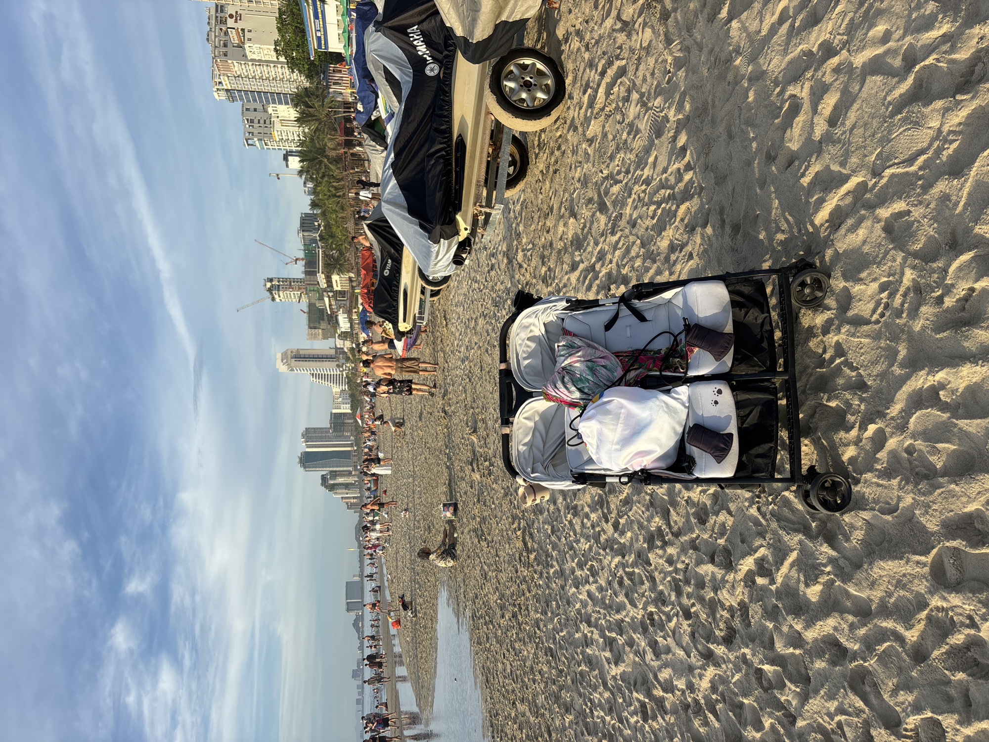 This image depicts a busy beach scene in Da Nang, Vietnam, likely on My Khe Beach, characterized by its wide expanse of sand, shallow ocean waters, and a backdrop of modern city buildings and palm trees. In the foreground, a double stroller is parked on the sand, laden with various items, including a white garment and a patterned bag.
Several jet skis are parked on trailers behind the stroller, covered by tarps, with one tarp prominently displaying the "YAMAHA" logo. The beach is populated with numerous people engaged in leisure activities, with some wading in the shallow water and others walking along the shoreline.
In the mid-ground, a person is sitting on the sand near the water's edge with a bag, while other individuals are scattered across the beach. The background reveals a cityscape with high-rise buildings, suggesting a well-developed urban area adjacent to the beach. The sky is a bright blue with scattered white clouds, and construction cranes are visible amongst the buildings, indicating ongoing development in the city. The overall atmosphere appears to be that of a popular tourist destination during daylight hours.