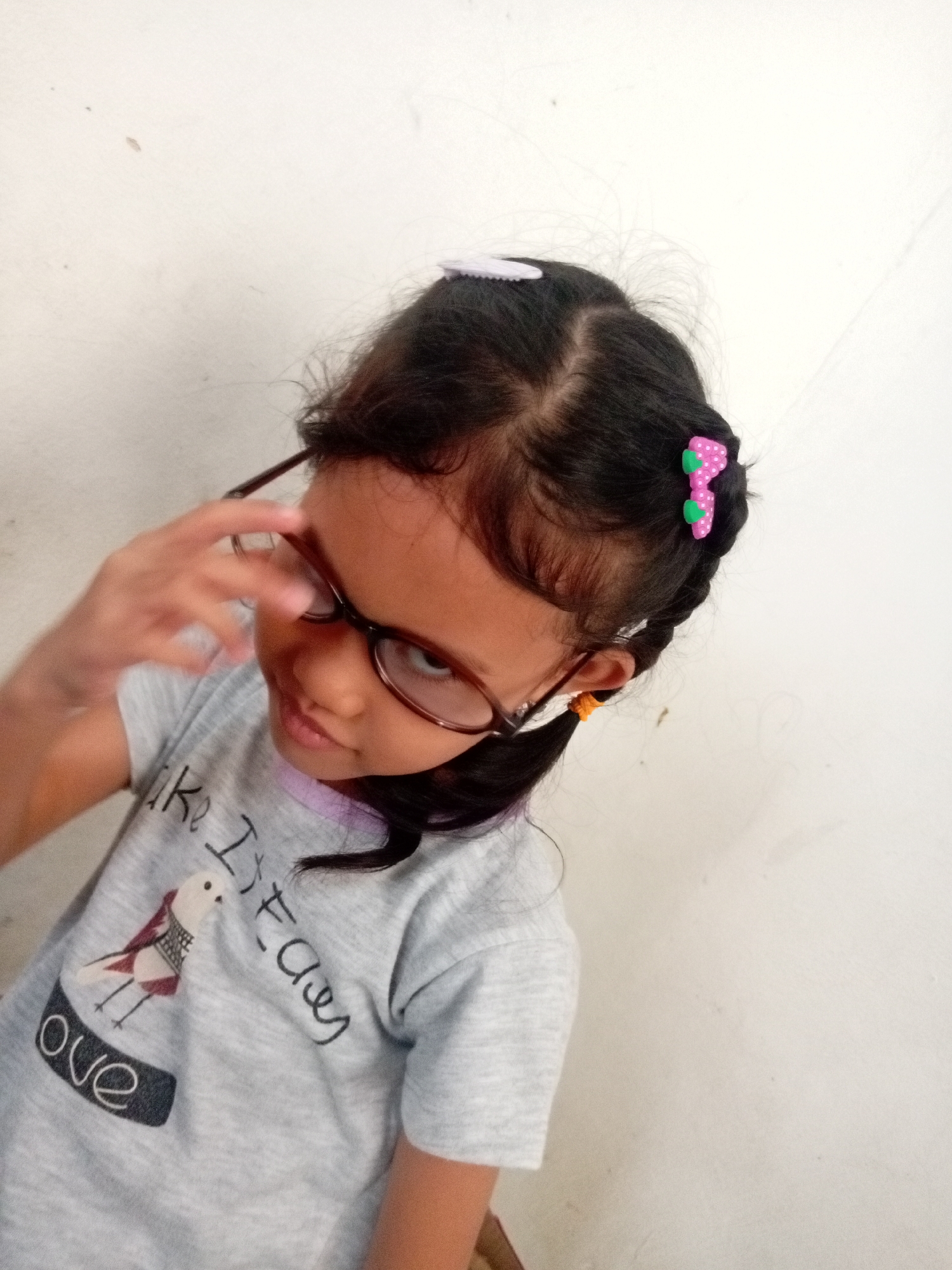 A young Indonesian girl with dark hair, glasses, and a grey t-shirt stands against a plain white wall. She is wearing a pink polka dot hair clip and has a braid. Her t-shirt has a graphic of a bird and the word "ove". The image was taken indoors, possibly in a home in Batam, Indonesia.