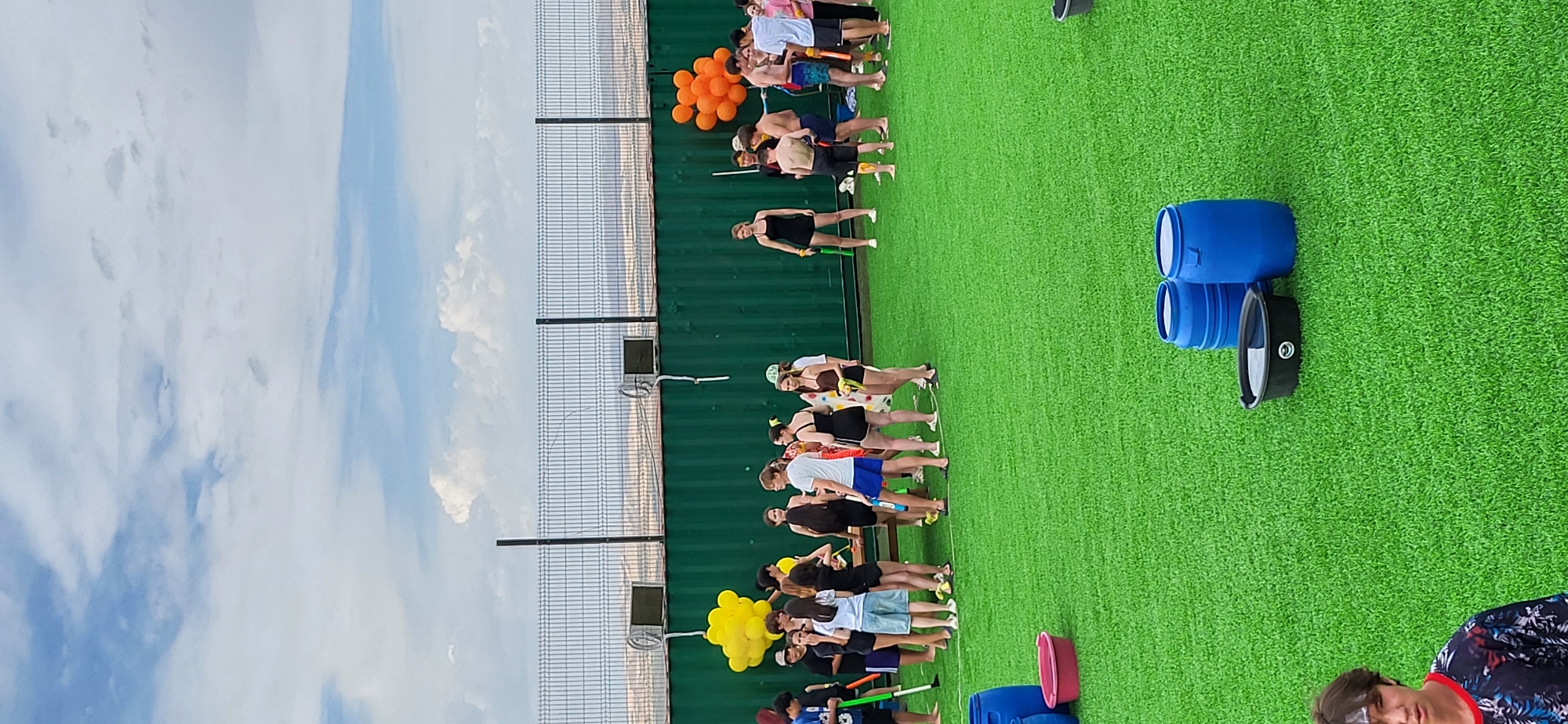 In the city of Syrymbet, Kazakhstan, an outdoor gathering takes place on a bright, overcast day. A group of young people, predominantly teenagers and young adults, are positioned on a vibrant green artificial turf surface. Some are standing in clusters, others are more spread out. Many are holding water guns, suggesting a water-based activity or game. A long, dark green container structure forms a backdrop, with a chain-link fence and distant hills visible beyond it. Yellow and orange balloons are clustered to the left and right, respectively, adding a festive touch. Several blue barrels and a black tub filled with water are scattered across the turf in the foreground, indicating preparations for or the aftermath of a water fight. The overall atmosphere appears casual and recreational.