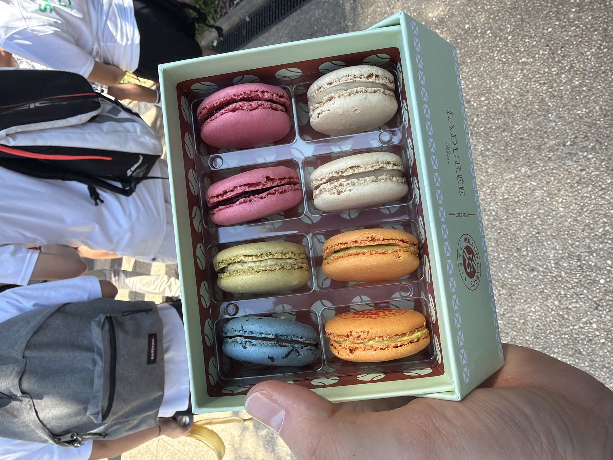 The image depicts a hand holding a light green Ladurée macaron box, customized for Roland Garros. The box contains eight macarons in different colors: pink, white, yellow, blue, and orange. The macarons are arranged in two rows of four inside a clear plastic container. In the background, other people are visible, wearing white shirts and carrying backpacks. The scene is likely in Paris, France, at Roland Garros or a nearby location.
