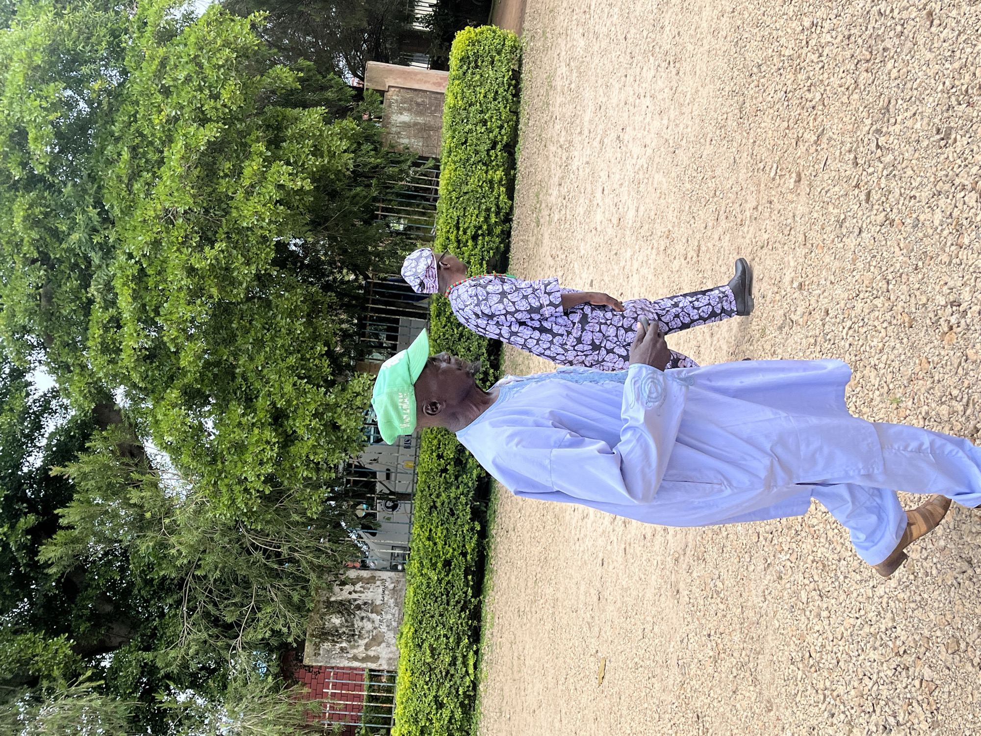 In the city of Vom, Nigeria, two individuals are captured in a side-profile, medium-shot, walking outdoors on a gravel path. The person in the foreground, an elderly man, is wearing a light blue traditional garment and a green cap. To his right, a person in a purple and white patterned traditional outfit walks in the same direction. Behind them, a dense green hedge forms a boundary, with trees and a building structure visible in the background, suggesting an urban or semi-urban environment. The lighting indicates an overcast day.