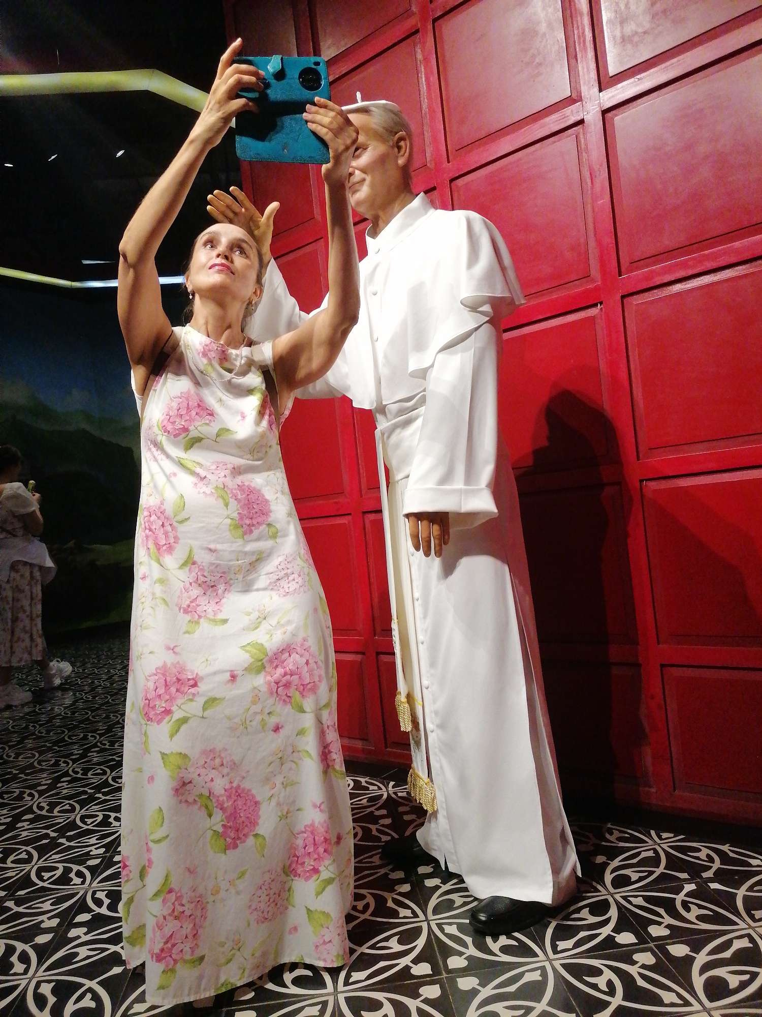 A woman in a floral sundress is taking a selfie with a wax figure of Pope John Paul II. She is holding up a blue tablet to take the picture. The location is in Hong Gai Ward, Vietnam. The background includes a red wall and a patterned black and white floor. There is a person in the background to the left.