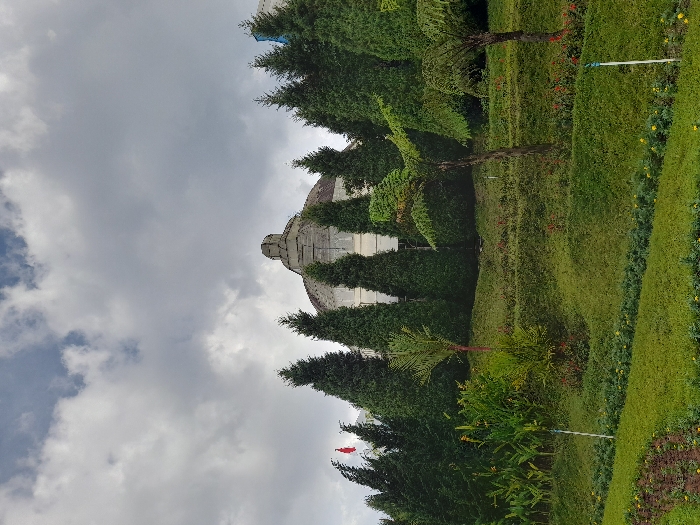 A large, grey stone building with a dome is partially obscured by tall, green evergreen trees. There is a small, red flag flying atop one of the trees. The building is located at the top of a grassy, terraced hill. The hill is covered in green grass and small green bushes. In the foreground, there are more evergreen trees and tall green plants. There is a clear, blue sky with white clouds. The scene is located in Kecamatan Pujon, Indonesia.