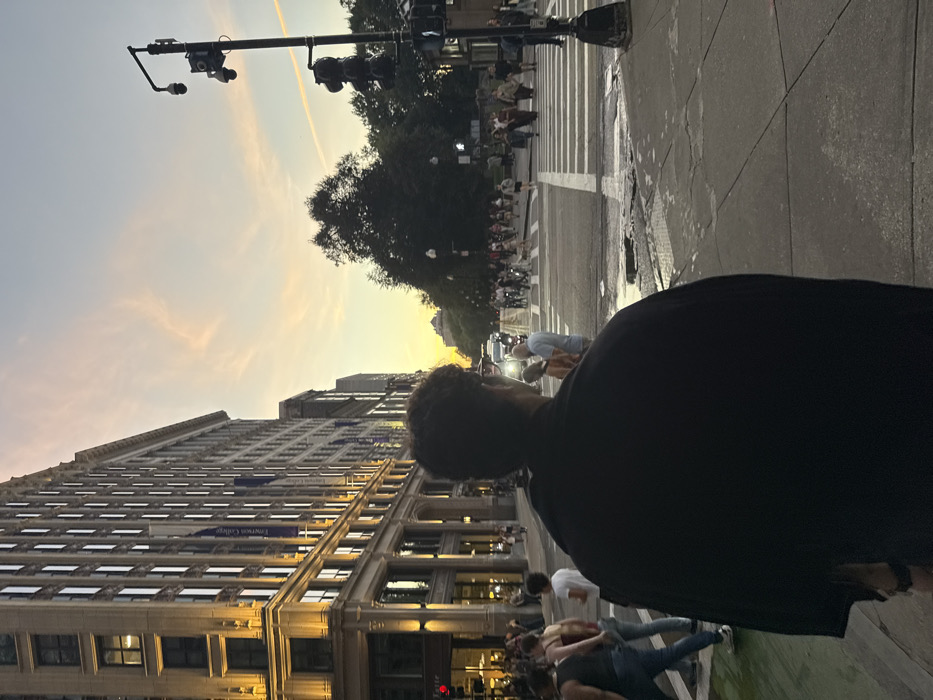 The image depicts a dusk scene at a city intersection in Boston, Massachusetts, United States. In the foreground, a person with dark, curly hair, viewed from behind, stands on a concrete sidewalk wearing a dark, possibly black, garment. To the left, a tall, multi-story building with "Emerson College" banners displayed vertically on its facade is brightly lit on its lower levels. The street in the midground features multiple lanes, white crosswalk markings, and several puddles on the asphalt surface. A green-painted bike lane is visible on the far left. Numerous pedestrians are either crossing the street, waiting on sidewalks, or congregated near the Emerson College building. Several vehicles with illuminated headlights are present on the street. On the right, a tall black pole supports traffic lights and at least two surveillance cameras. Beyond the street, a dense cluster of trees signifies a park area, likely Boston Common. The sky exhibits sunset colors, transitioning from orange and pink near the horizon to light blue above, with faint clouds and a distinct contrail.