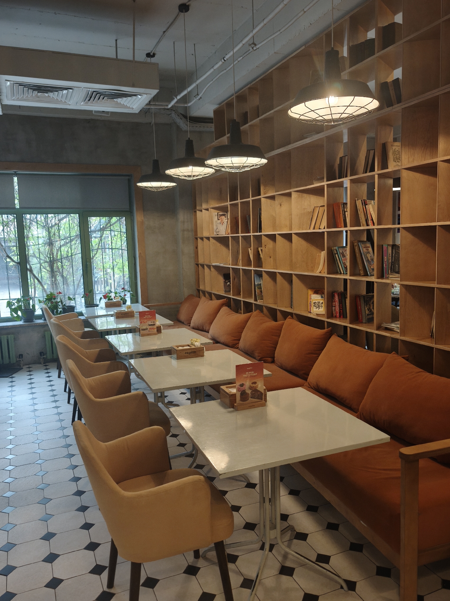 A well-lit interior view reveals a cafe or restaurant setting in Unknown, Unknown. The floor is covered in a tessellated pattern of black and white tiles. Along one wall is a built-in wooden bookcase filled with books and various items, alongside an orange cushioned bench seat. Three light fixtures hang from the ceiling, emitting a bright glow. Four square white tables with metal legs are positioned in front of the bench, each accompanied by several beige upholstered chairs. A large window allows natural light to filter into the space, and potted plants are placed along the windowsill. The walls are a neutral gray color.