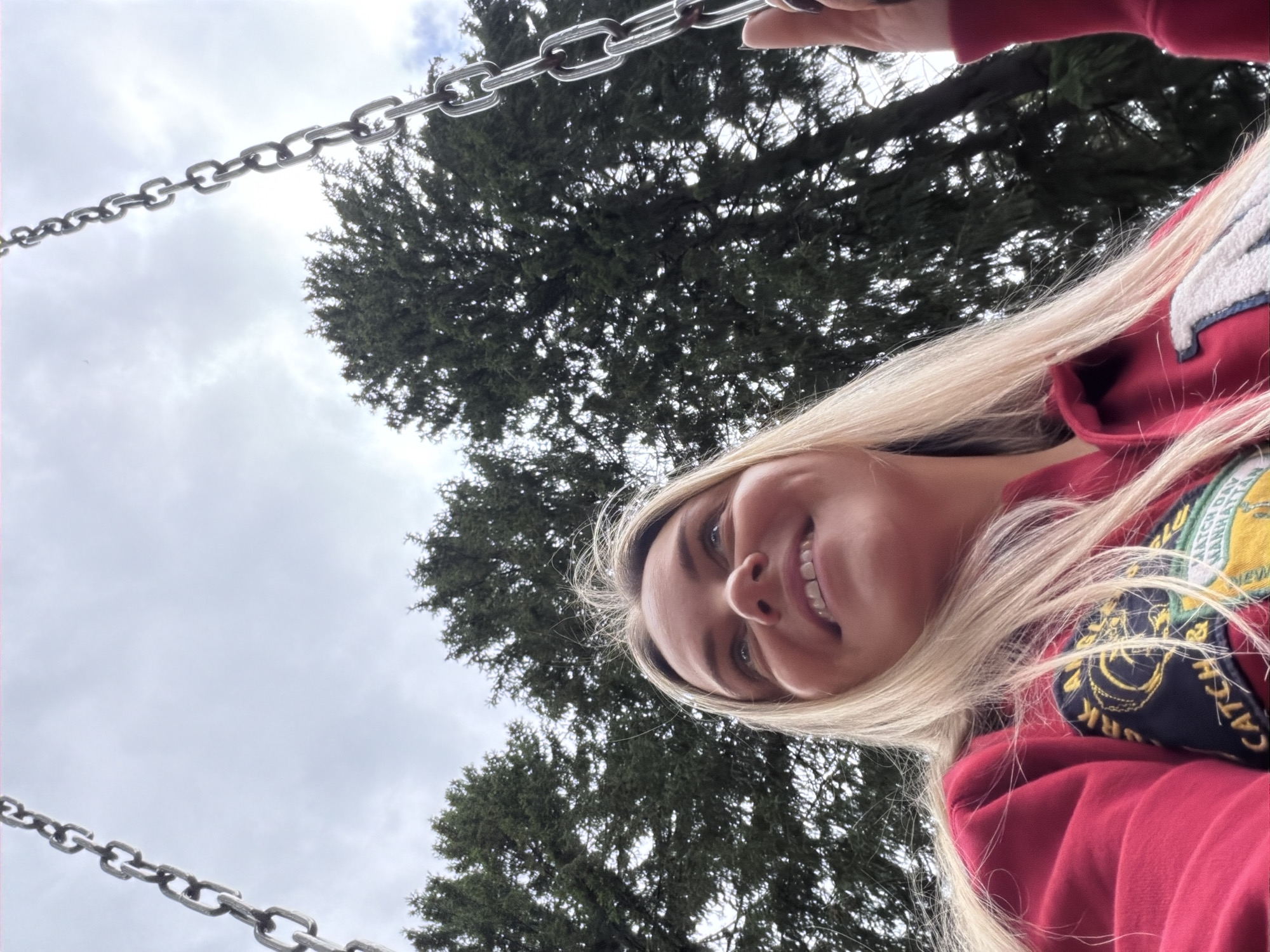 A young woman with blonde hair is pictured from a low angle, looking up. She is wearing a red hooded sweatshirt with patches on the chest. She is on a swing, indicated by the metal chains visible in the frame. Tall evergreen trees are in the background, and the sky is overcast. The scene appears to be outdoors, possibly in a park or backyard. There are no discernible features that place this image specifically in Votkinsk, Russia; it is a general outdoor scene.
