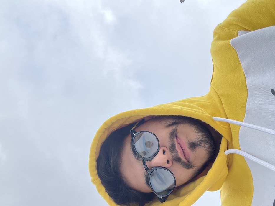 The image depicts a young adult male, positioned at a low angle and looking directly into the camera against an overcast sky. He has dark hair, a beard, and a mustache, and is wearing round-framed black sunglasses. Visible dimples are present on his cheeks. He is dressed in a yellow hooded sweatshirt with a prominent white horizontal stripe across the chest and white drawstrings. The hood is pulled up, framing his head. The background consists predominantly of a light grey, cloudy sky. A faint, indistinct, light-colored structure or object is minimally visible on the far right edge of the frame against the sky. The scene is located in Al-Muhajerin Municipality, Syria.