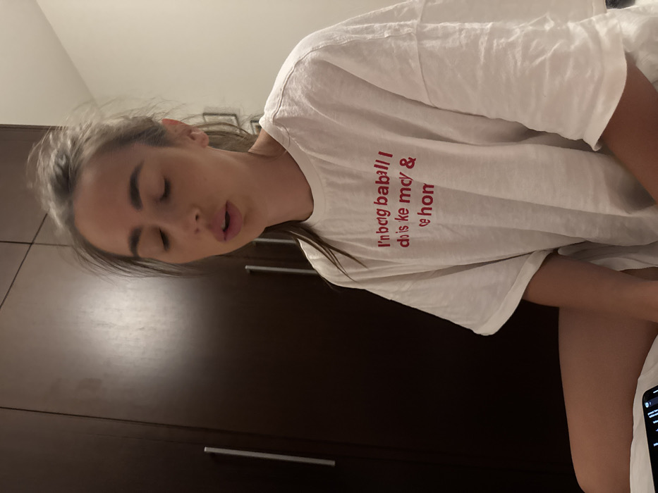 A woman with closed eyes, possibly sleeping, is photographed indoors. She wears a white t-shirt with red text that reads, "I'm bag baball I dois ke mcy & te hon." A dark brown wardrobe with metal handles is visible in the background. A smartphone is partially seen in the lower right corner of the image. The overall impression is of a casual, unposed moment.