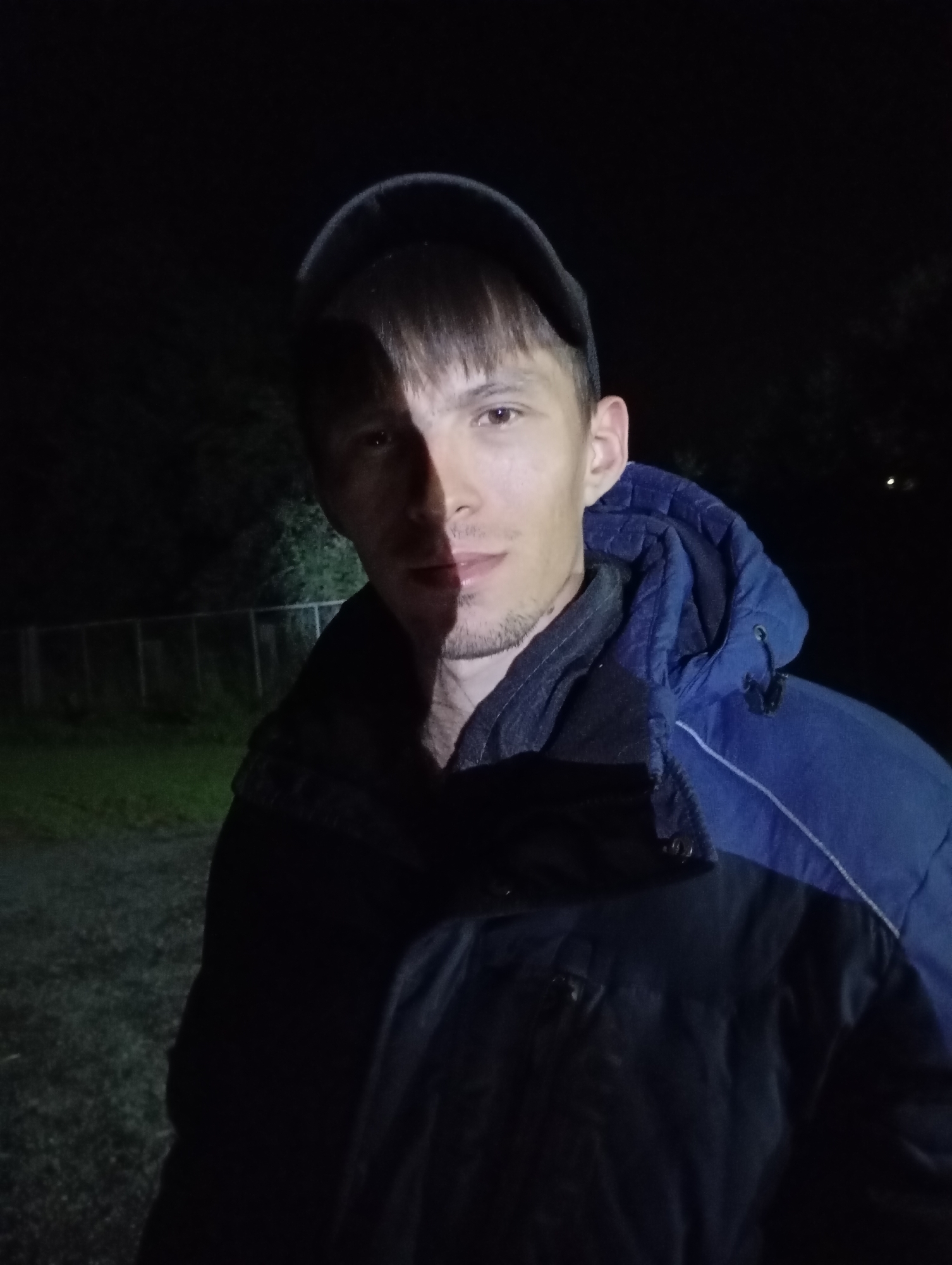 A medium shot, taken at night, shows a person from the chest up, facing forward. The person is wearing a black baseball cap, a dark jacket over a black shirt, and a blue hooded jacket. A light source illuminates the left side of the person's face, casting the right side into shadow. The background is dark, with some greenery and a fence visible to the left. This scene is located in the city of Malyi Bukor, Russia.