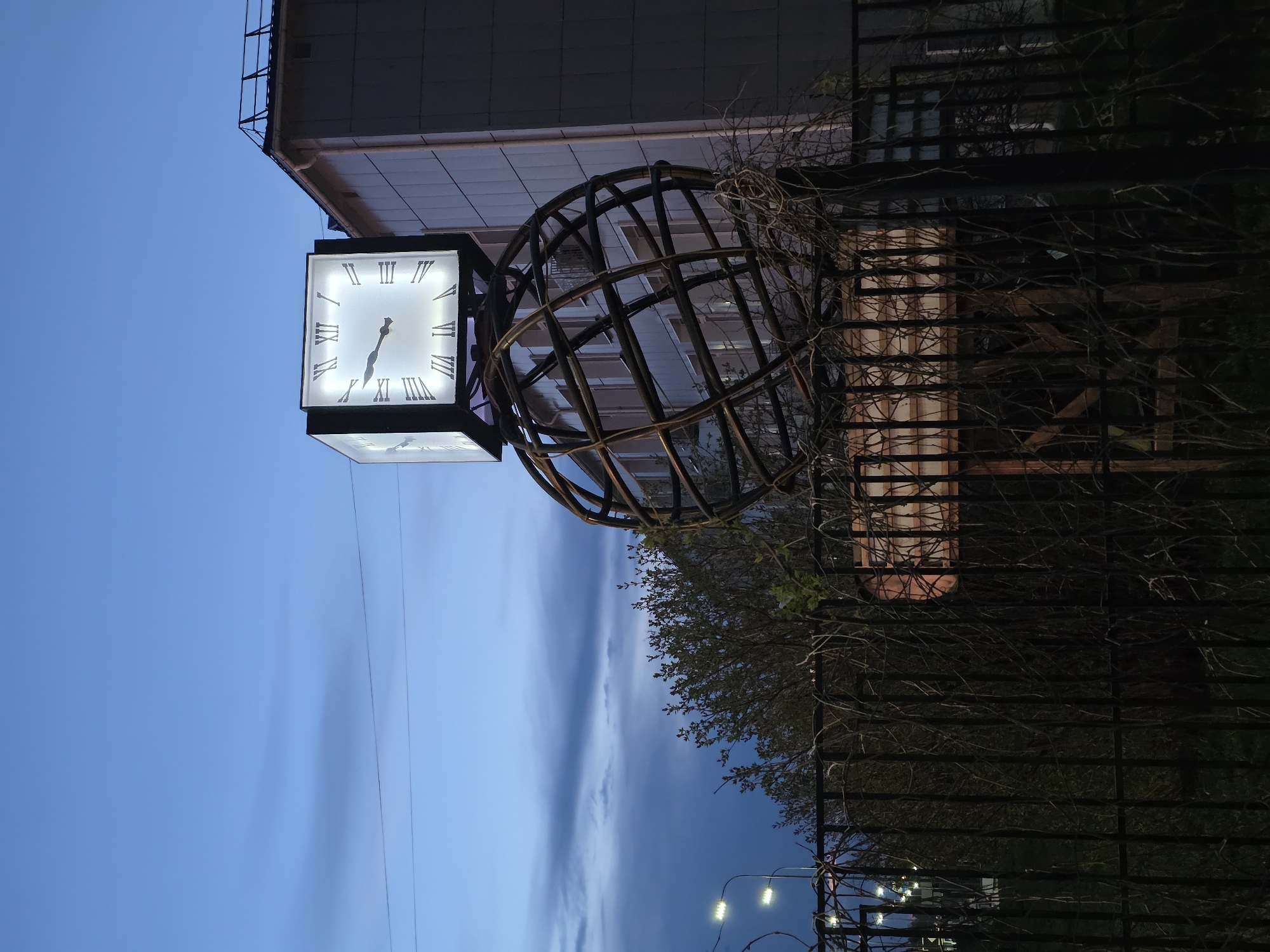 A street scene in Chaikovsky, Russia, shows a large square clock with Roman numerals at about 6:25. The clock sits on a metal stand with a sphere design. A building is in the background, and a fence runs along the right side. There are trees and bushes, and the sky is a dusk blue. Streetlights are visible in the bottom right corner.