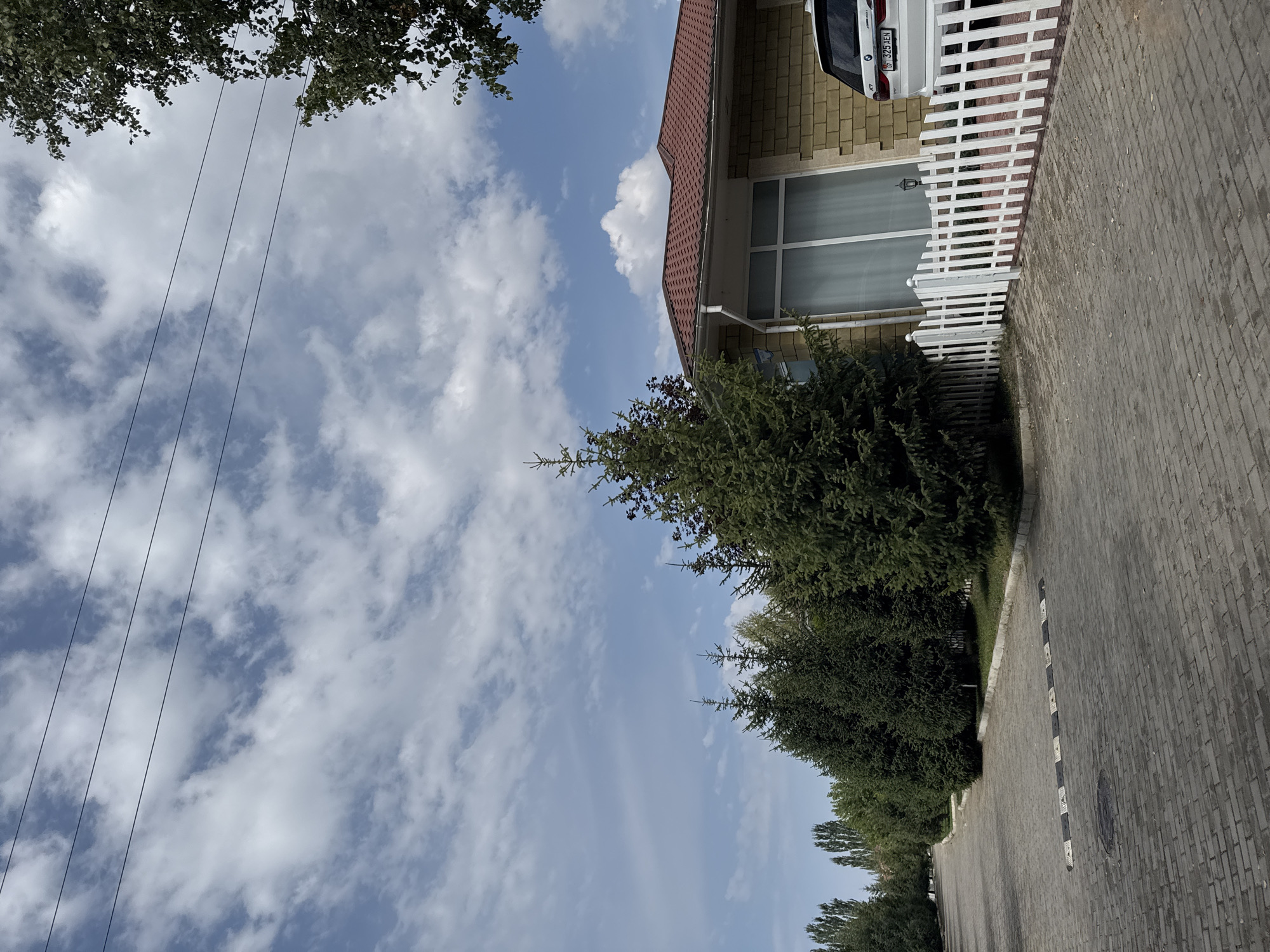 The image shows a paved road in the city of Sary-Oy, Kyrgyzstan. A line of coniferous trees runs along the left side of the road, with a few deciduous trees further in the distance. On the right side of the road, there is a house with a red tiled roof and yellow brick siding. A white picket fence encloses the front yard of the house, and a white SUV is parked in the driveway. The sky is partly cloudy with a few scattered white clouds against a blue background. There are three visible overhead power lines stretching across the upper portion of the image.