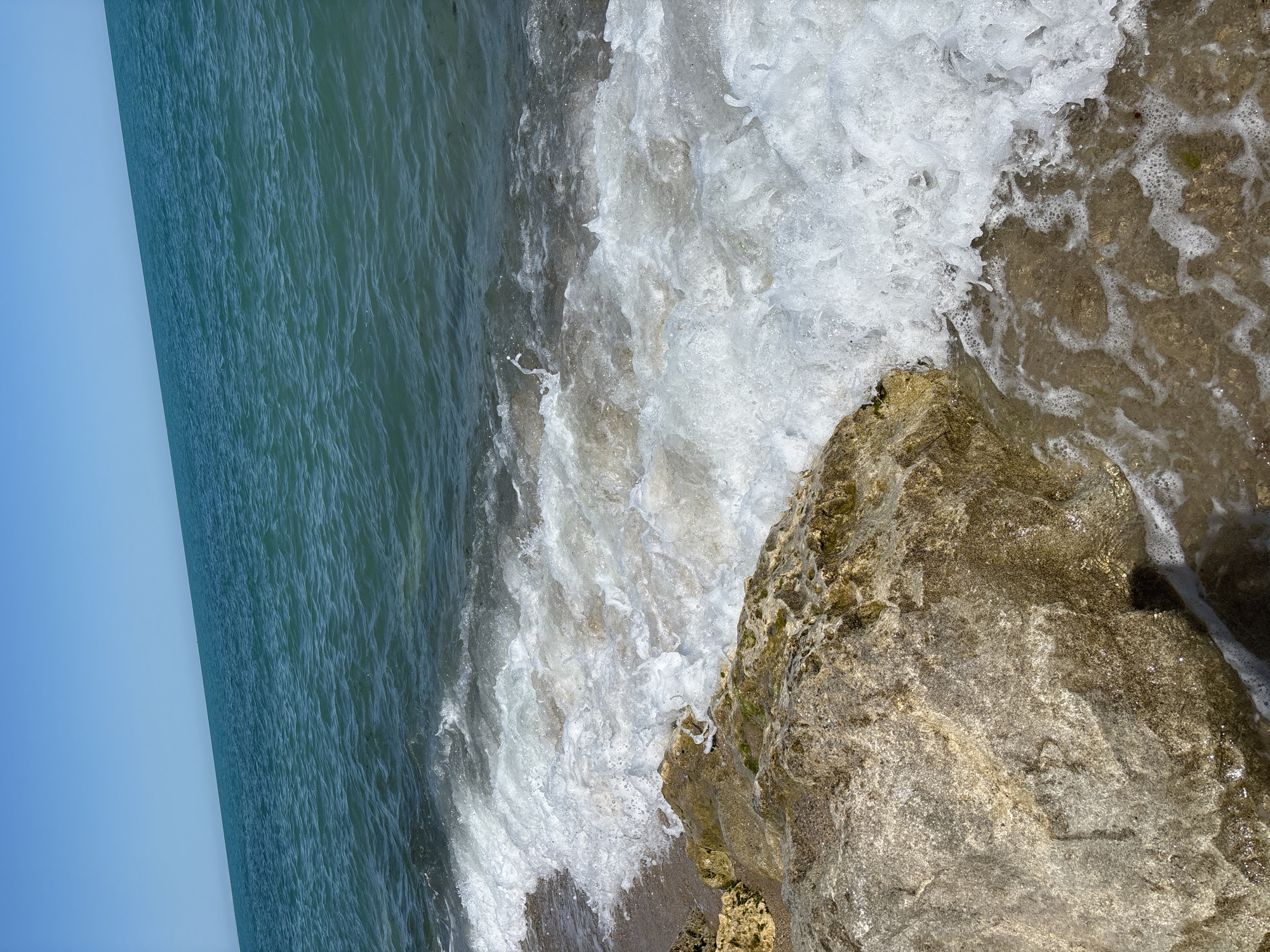 The image depicts a close-up view of the Black Sea coast near Varna, Bulgaria, on a clear, sunny day. The foreground is dominated by rough, textured rocks, some of which are wet and show patches of green algae. Waves are actively crashing onto the shore, creating white foam and churning the shallow water. The sand beneath the water is visible and appears wet. The sea itself is a deep turquoise color, with ripples and gentle waves extending towards the horizon. The horizon line is sharp, dividing the sea from a pale blue, cloudless sky. No people or man-made structures are visible in the frame.