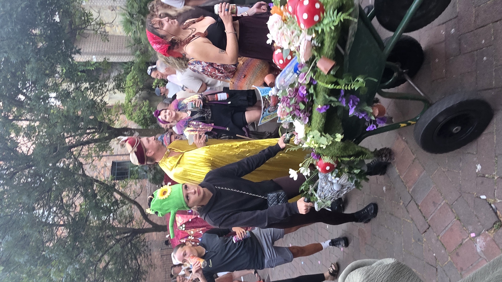 The image captures a street scene in St Leonards, United Kingdom, possibly during a parade or festival. In the foreground, a young woman wearing a black hoodie and leggings pushes a wheelbarrow adorned with greenery, purple flowers, and red toadstools. She wears a green hat shaped like a mushroom with a yellow sunflower on it. To her left, another young woman in a grey athletic shirt and shorts holds a can. Behind them, a man in a bright yellow robe and a red and gold hat is visible, along with other festival-goers in eclectic attire. The background features brick buildings and trees, suggesting an urban outdoor setting. The ground is paved with cobblestones.
