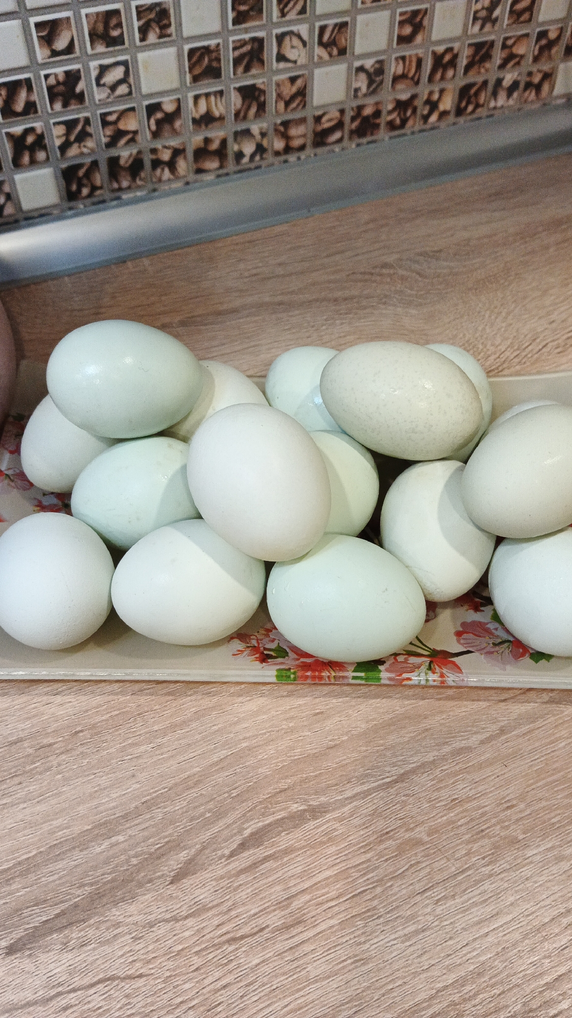 A pile of pale blue-green eggs is displayed on a clear tray with a floral pattern. The tray rests on a wooden surface. In the background, a tiled wall is visible above a silver countertop. The scene is well-lit, suggesting it might be indoors. There is no indication of the city of Kryvyi Rih, Ukraine in this image.