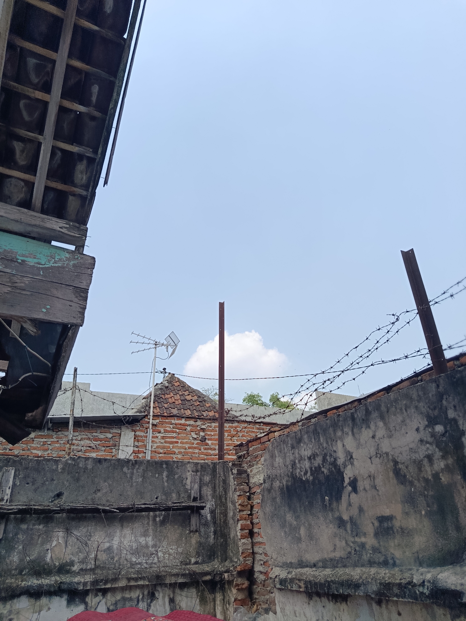The image depicts a partially constructed or dilapidated building, likely in an urban setting within the city of Madiun, Indonesia, given the characteristic roofing tiles and brickwork. The foreground is dominated by a rough, weathered concrete wall, showing signs of age and damage, with sections of exposed brickwork and crude wooden supports. Barbed wire fences are strung across the top of the wall and on metal posts, suggesting an attempt at security or boundary marking.
In the mid-ground, a similar brick structure with a tiled roof is visible, adorned with an antenna. The architectural style appears consistent with older buildings in Indonesia. A reddish-brown metal pole stands upright behind this structure. Above the buildings, a pale blue sky stretches across the frame, with a few wispy white clouds present.
The left side of the image features a section of an overhanging wooden roof structure, made of dark, weathered planks supported by exposed beams, indicating a state of disrepair. The overall impression is one of an unfinished or decaying urban environment, possibly an industrial area or a residential neighborhood undergoing development or experiencing neglect. There are no people or animals visible in the frame. The lighting suggests it is daytime, with natural light illuminating the scene.