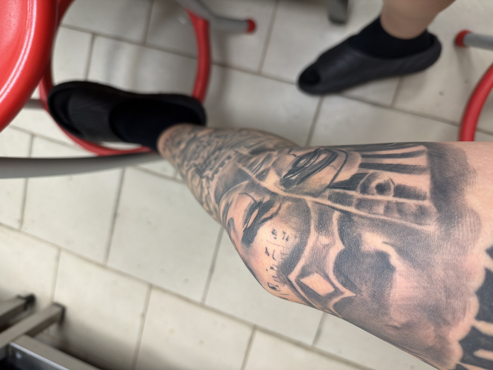 The image shows a close-up view of a person's leg, covered in extensive black and grey tattoos, resting on a red object that appears to be part of a chair or play structure. The person is wearing black socks and black slide sandals. Another person's leg, also wearing a black slide sandal and black sock, is visible in the background, suggesting a shared space. The floor is tiled with light-colored square tiles. The scene appears to be indoors, possibly in a public or recreational area, given the style of the furniture and flooring. There is no discernible text in the image. The lighting suggests daytime. The overall mood is casual and relaxed.