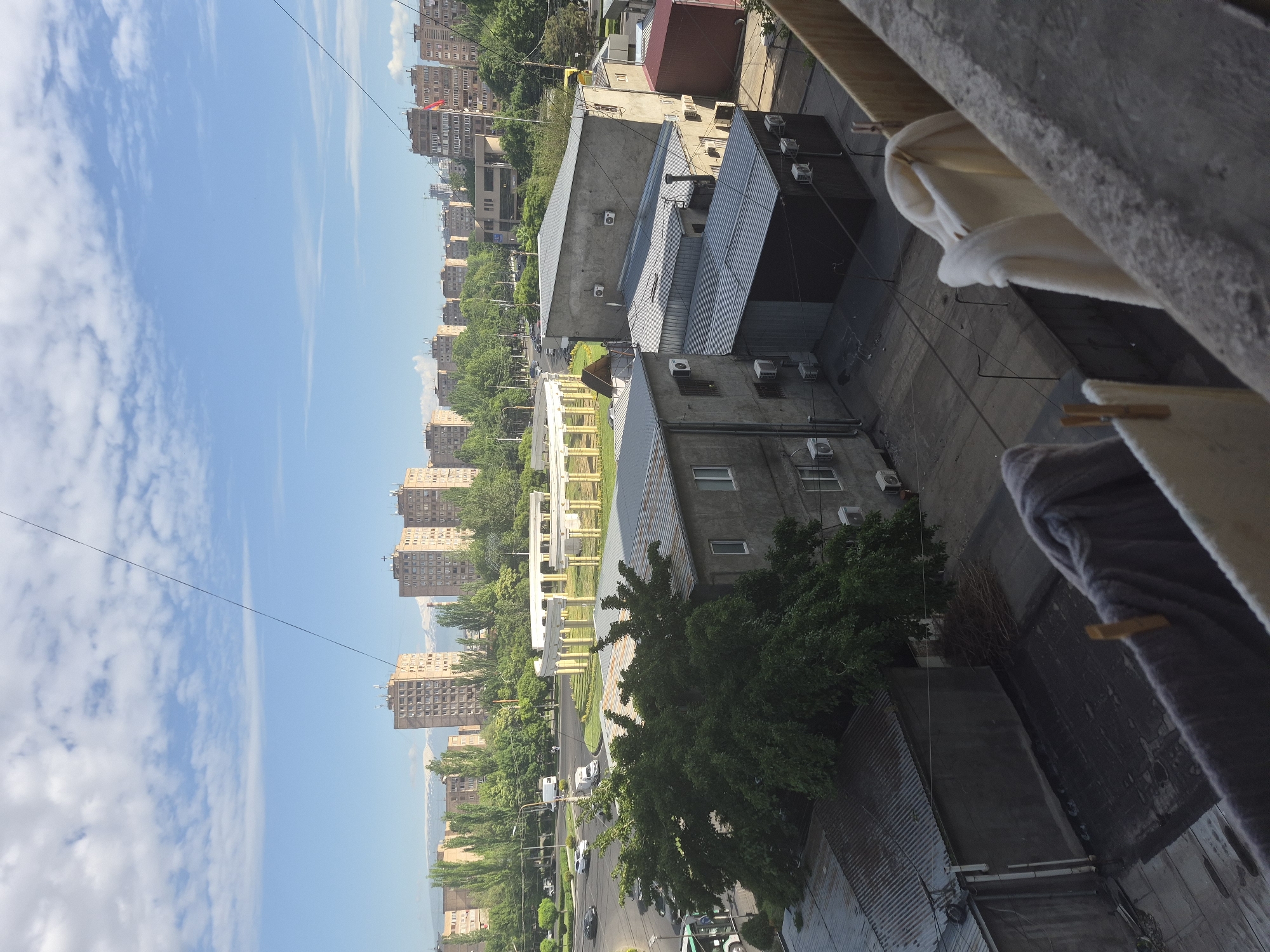 The image captures a panoramic view of an urban landscape in Yerevan, Armenia, from a high vantage point, possibly a balcony. In the immediate foreground, a concrete railing is visible, over which white and grey/purple towels or blankets are hanging to dry, secured with wooden clothespins.
Below and to the right, a series of low-rise, utilitarian buildings with corrugated metal roofs and concrete walls dominate the midground. Numerous air conditioning units are mounted on their facades. Interspersed among these structures are scattered green trees.
Further into the scene, a wide, tree-lined street stretches horizontally across the image, with several cars, including a white van, parked or moving. Alongside the street, a pedestrian area features decorative white or light-colored structures, possibly arches or columns. Beyond this, rows of tall, multi-story apartment buildings, characteristic of Soviet-era architecture, extend into the distance, partially obscured by lush green trees.
In the far background, faint outlines of mountains are visible beneath a clear, bright blue sky dotted with scattered wispy white clouds. The bright, even lighting suggests it is daytime, likely morning or early afternoon, with sunny and clear weather. The scene depicts a typical urban environment, focusing on architecture, street life, and domestic activities like laundry, with no discernible people or visible text.