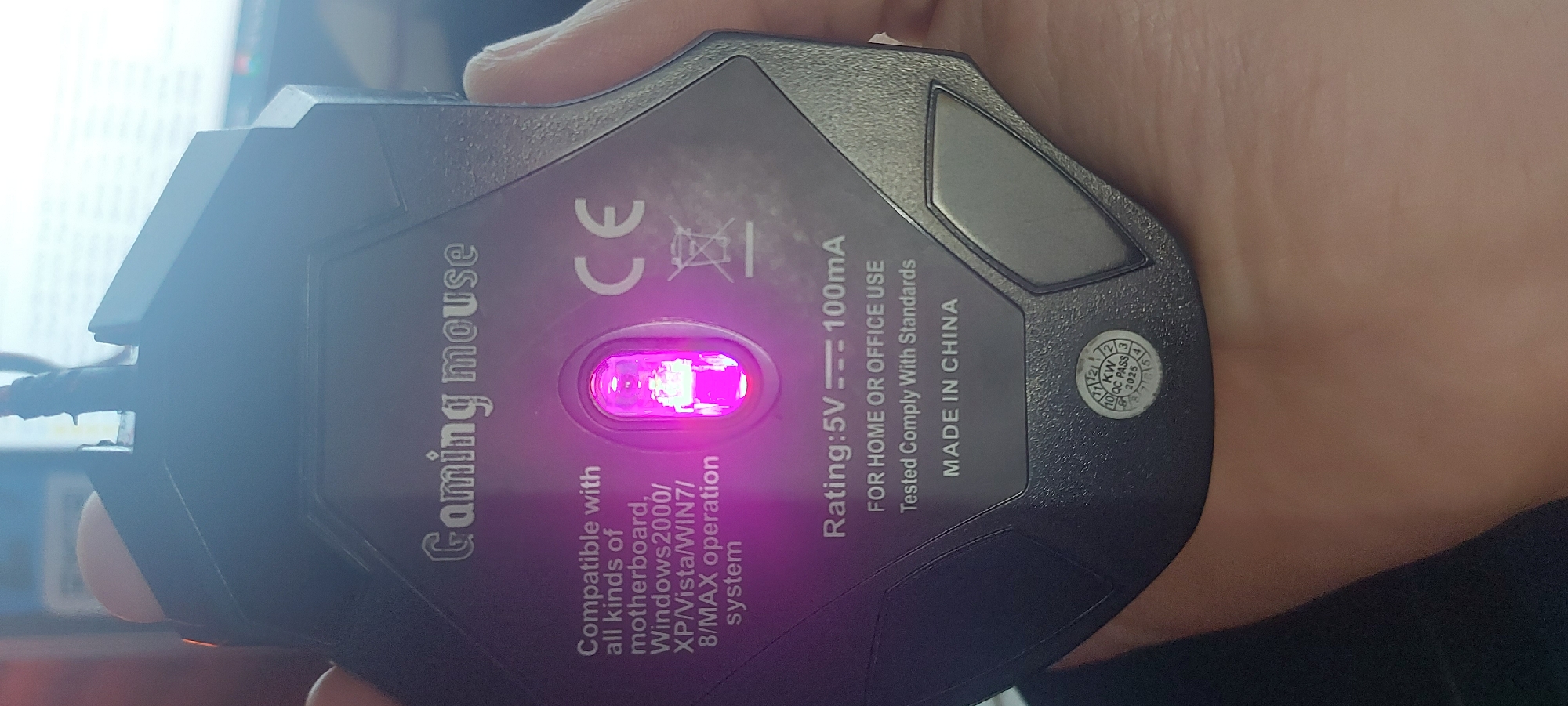 The image captures the underside of a black gaming mouse being held by a person's hand in an indoor setting. The mouse features a bright, glowing magenta optical sensor at its center. The surface of the mouse has a subtle texture, and a section of its attached cable is visible at the top left of the frame. The background is brightly lit and blurred, suggesting a computer monitor or another light source in a workspace.
The underside of the mouse displays several labels and specifications. Prominently visible is the text "Gaming mouse." Further details include "Compatible with all kinds of motherboard, Windows2000/ XP/Vista/WIN7/ 8/MAX operation system." The "CE" conformity mark is present, along with a crossed-out wheeled bin symbol, indicating proper disposal guidelines. Electrical ratings are specified as "Rating: 5V --- 100mA." Other informational text states "FOR HOME OR OFFICE USE," "Tested Comply With Standards," and "MADE IN CHINA." A circular "KW QC PASS 2025" sticker is affixed, with an indicator pointing to the number '3'. The hand holding the mouse is light-skinned. The scene is likely a desk environment in Tashkent, Uzbekistan.