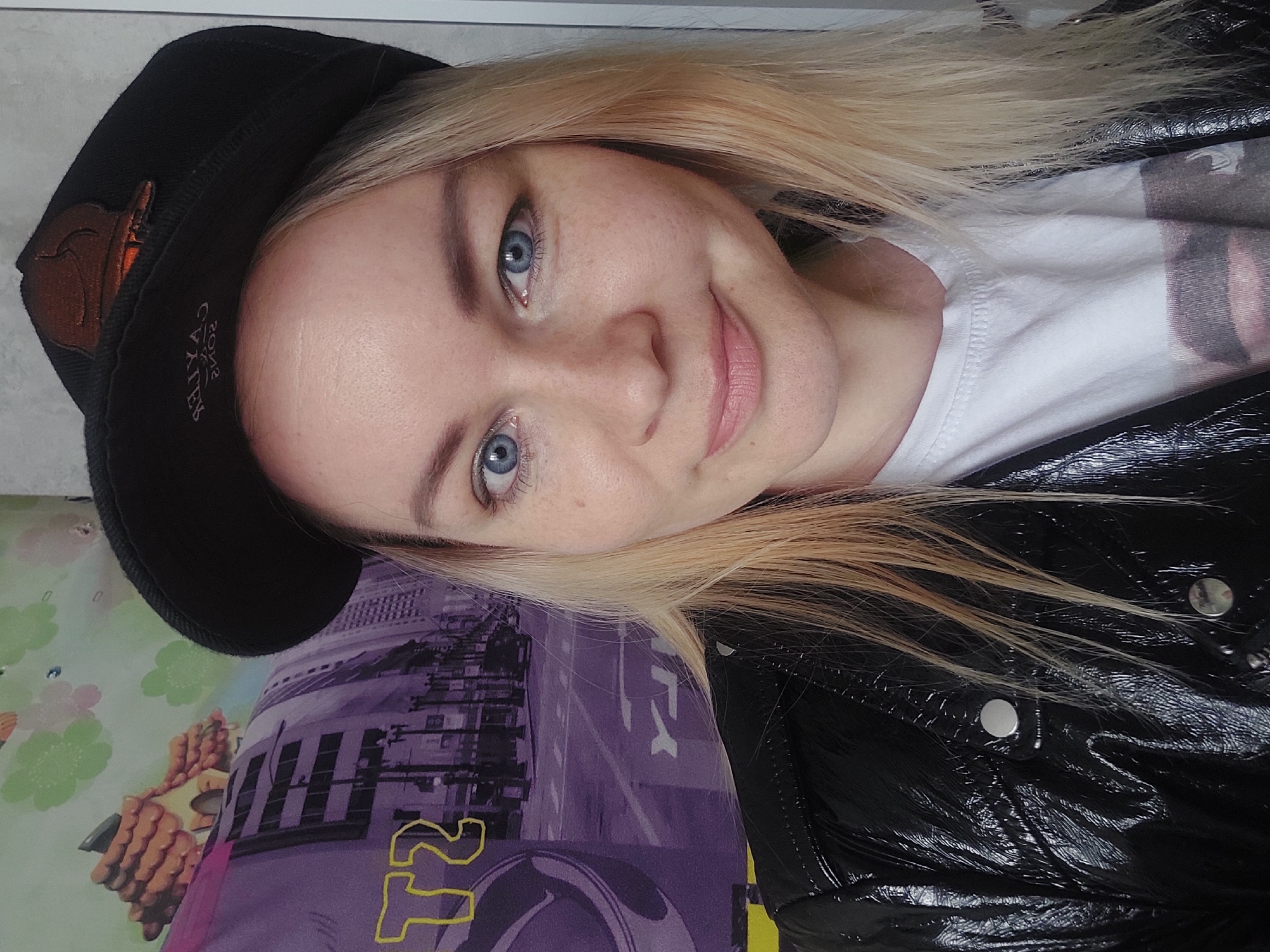 A young woman with blonde hair and blue eyes is facing the camera, wearing a black cap with an orange patch, a white t-shirt, and a shiny black jacket. The background features a purple pattern with depictions of urban buildings, possibly indicating a scene from a city. The presence of what looks like a cityscape design could potentially point to a location with urban architecture. The context of Chaikovsky, Russia is not represented.