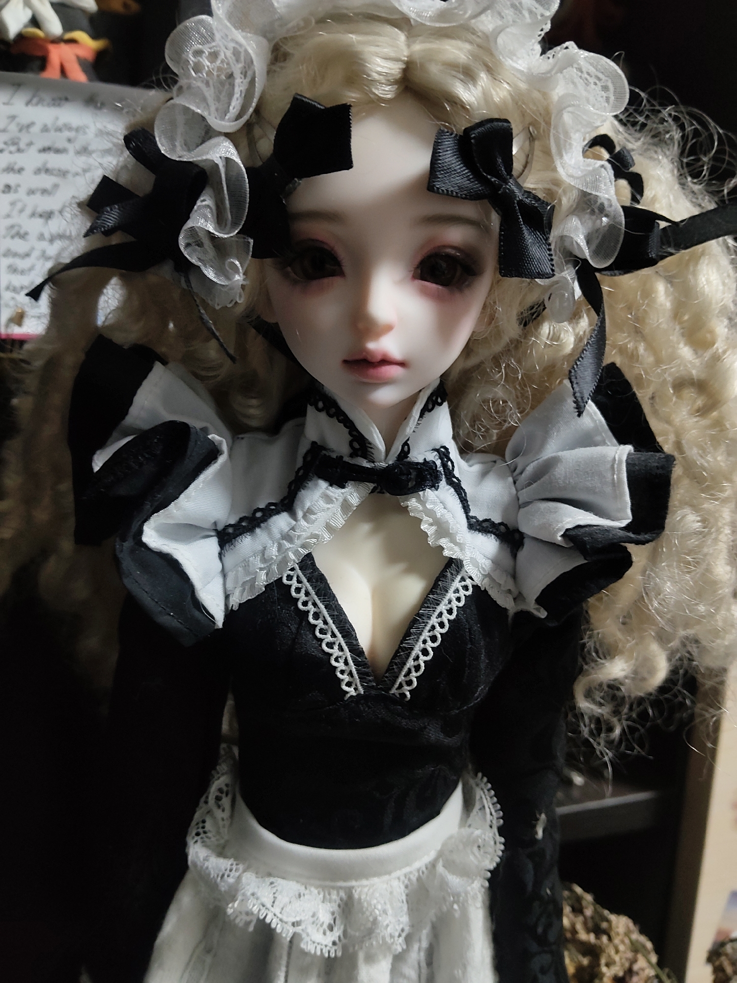 The image shows a close-up of a doll with blonde curly hair and fair skin. The doll is wearing a black and white outfit with lace trim and black bows in its hair. The background is blurred and appears to be an indoor setting with some papers and objects visible. The image is a photograph and does not provide any information about the location in Kharkiv, Ukraine.