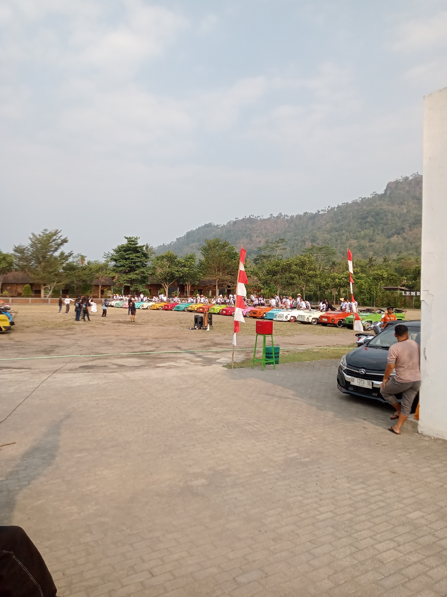 The image depicts an outdoor gathering, likely an event, in Majaksingi, Indonesia. In the foreground, a gray car with license plate "AB 1373 XK" is parked near a white building. A person wearing a light brown t-shirt and gray shorts is leaning against the white building, with their back to the camera, looking towards the car. To the left of the car, a person in a dark t-shirt and dark pants is visible from the waist down, partially obscured by the building.
The middle ground reveals a large open area, possibly a field or parking lot, where a line of colorful miniature cars is parked. Red and white Indonesian flags are placed strategically near the miniature cars. A group of people, dressed in various casual attire, are gathered around the miniature cars, with some appearing to be participants or spectators. Behind the miniature cars, more people are visible, some standing and others seated. Trees and buildings with terracotta roofs are present in the background, suggesting a rural or semi-rural setting.
The far background is dominated by a large, tree-covered hill or mountain range under a hazy sky with scattered clouds. The overall impression is of a community event or festival taking place in a scenic, natural environment.