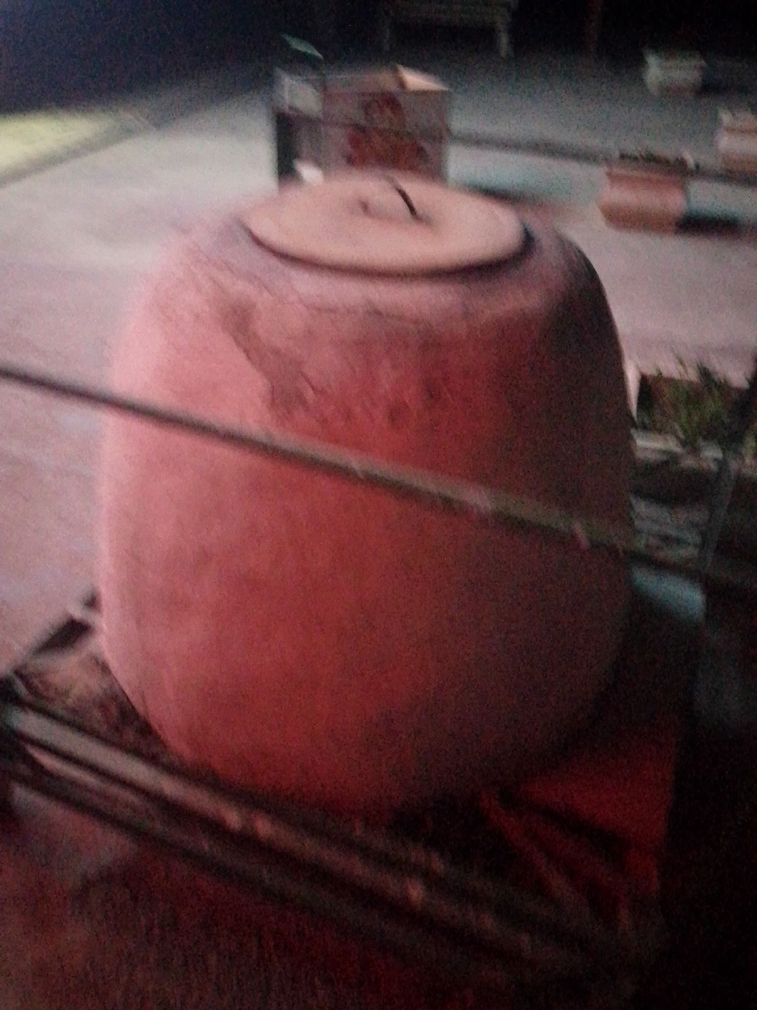 A large, reddish-brown, clay tandoor oven, with a lid slightly ajar, sits on a woven mat. The tandoor is the central and largest object in the frame, positioned in the foreground and slightly off-center to the left. It appears to be outdoors, with a paved surface visible behind it. In the background, out of focus, are several structures, possibly market stalls or buildings, and some greenery. A thin, dark, horizontal line, potentially a wire or a stick, runs across the front of the tandoor. The overall lighting is dim, suggesting it might be evening or a shaded area. No people are visible in the image. The scene appears to be a street or outdoor market in Bukhara, Uzbekistan, given the context of tandoor ovens being common in Central Asian cuisine.