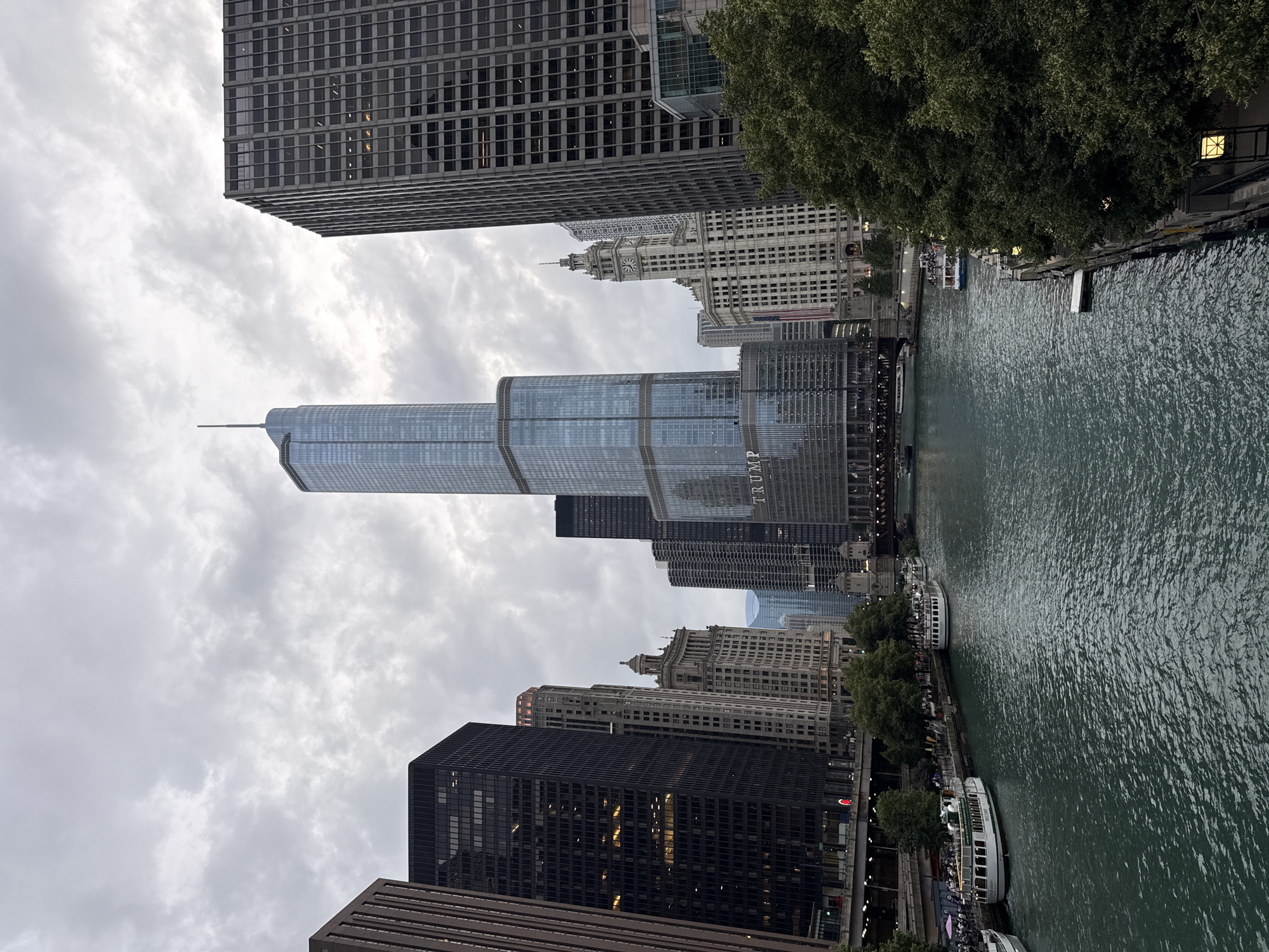 The image captures a view of the Chicago River and the city's skyline. In the foreground, the murky green water of the river flows. Along the riverbanks, several tour boats are docked or moving. On the left side, a white and green tour boat with "CITY LIGHTS" written on it is visible. Further down, another similar boat is docked. On the right bank, trees line the riverwalk, with a few boats visible further up.
Dominating the skyline are numerous skyscrapers. The most prominent structure is the Trump International Hotel and Tower, a tall, glass-covered building with a distinctive spire. To its left, another tall, slender skyscraper with a pointed top stands out. Various other buildings of different architectural styles and heights fill the background, creating a dense urban landscape. Some of the buildings have a grid-like facade, while others feature more decorative elements.
The sky is overcast with heavy clouds, casting a diffused light over the scene and giving it a somewhat subdued atmosphere. Bridges span the river in the distance, with traffic visible on them. The overall impression is that of a bustling city on a cloudy day, with the river acting as a central artery through the urban environment.
