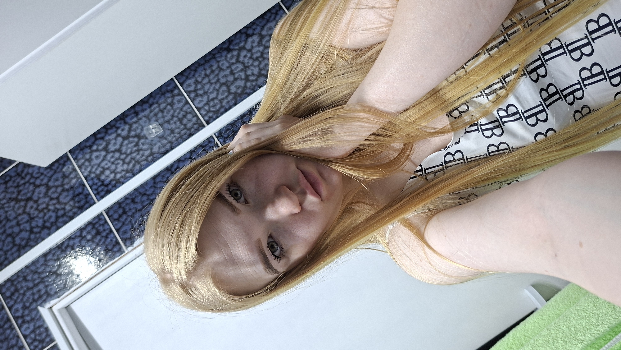 A selfie shot of a fair-skinned person with long, blonde hair is positioned in a bathroom. The person is wearing a white top with black "B" patterns. The background shows a bathroom wall covered in dark blue tiles with a patterned design and a white door or vanity. A green towel is visible in the lower right corner. The location is Chaikovsky, Russia.