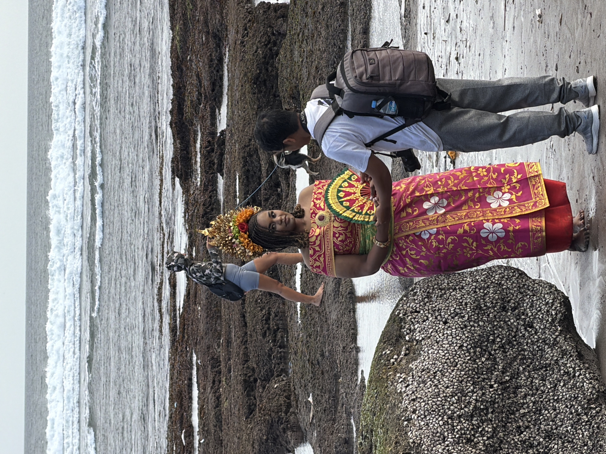 In the city of North Kuta, Indonesia, on a beach with dark, rocky shorelines and crashing waves in the background, a photographer is taking pictures of a woman dressed in traditional Balinese attire. The woman is wearing a vibrant pink and gold, off-the-shoulder outfit with a gold headdress adorned with flowers and a fan in her hands. The photographer, wearing a white t-shirt, grey pants, and a backpack, is holding a camera and appears to be interacting with the woman. In the background, another person with a backpack walks away from the shore.